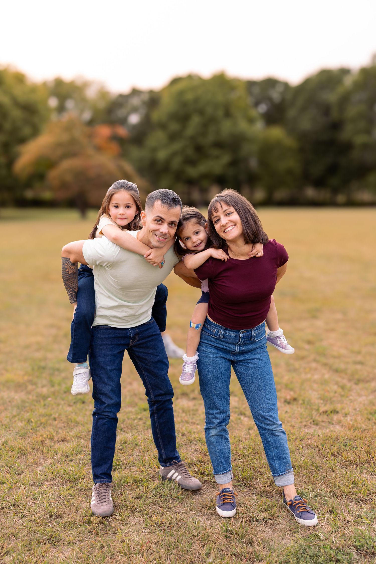 Happy family of four posing outdoors, parents giving piggyback rides to young daughters in a green park.