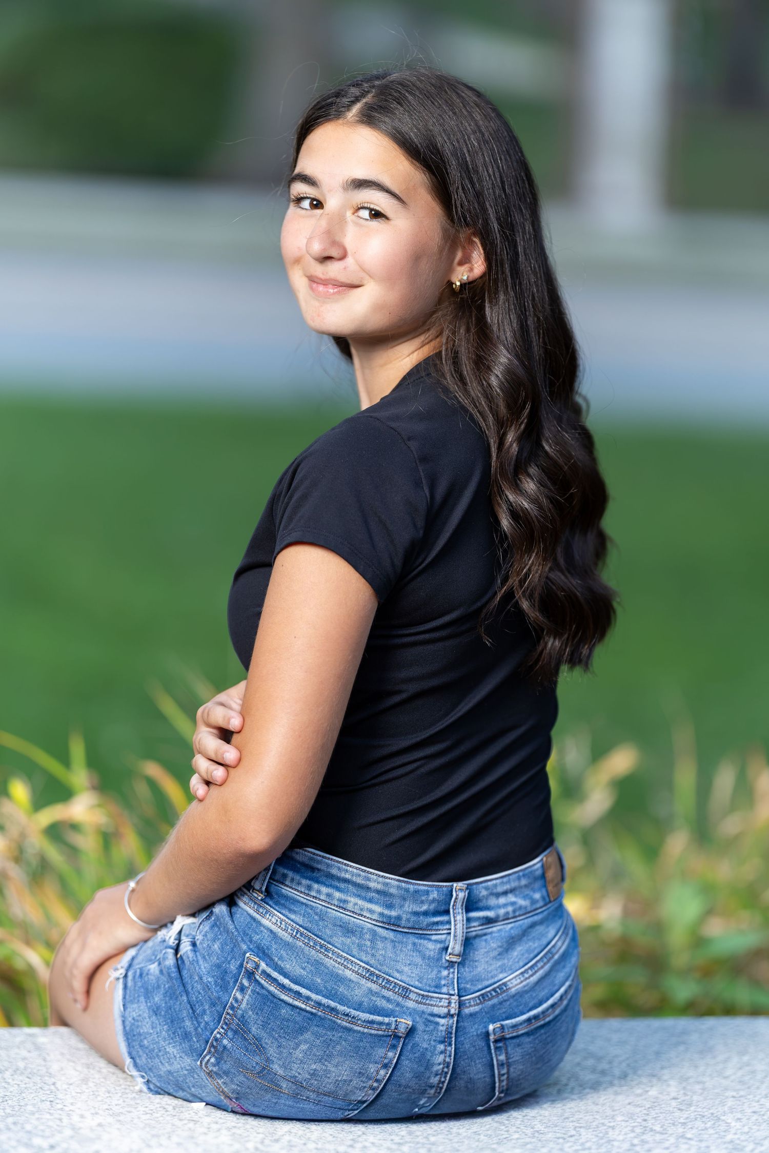 Teenage girl with long dark hair in black shirt and jeans sitting outdoors looking over her shoulder.