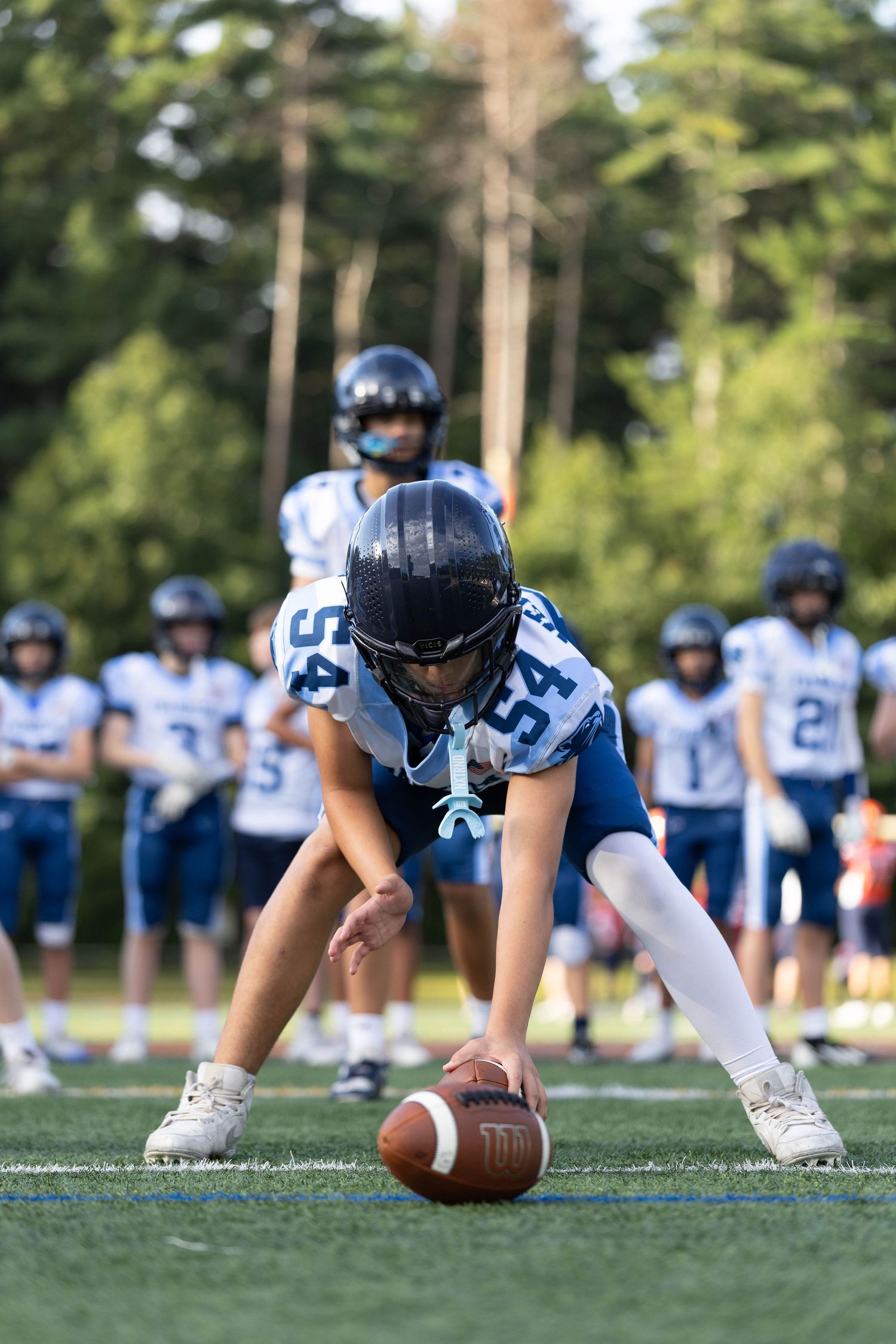 Young football player in blue and white uniform prepares to snap the ball during a sunny outdoor practice game.