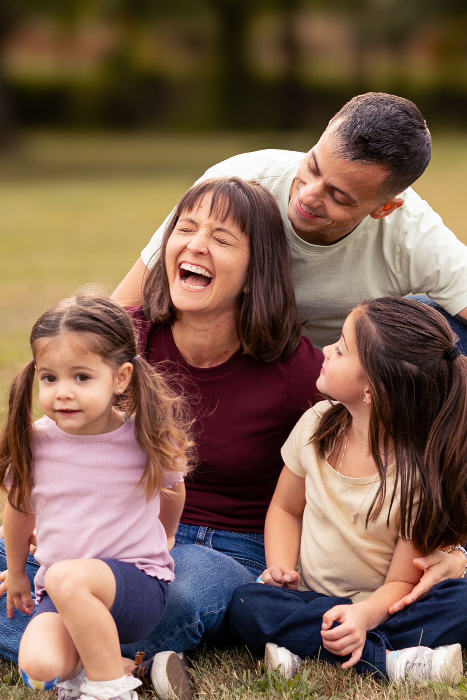 Joyful family of four sitting on grass in park, mother laughing while father and daughters smile.