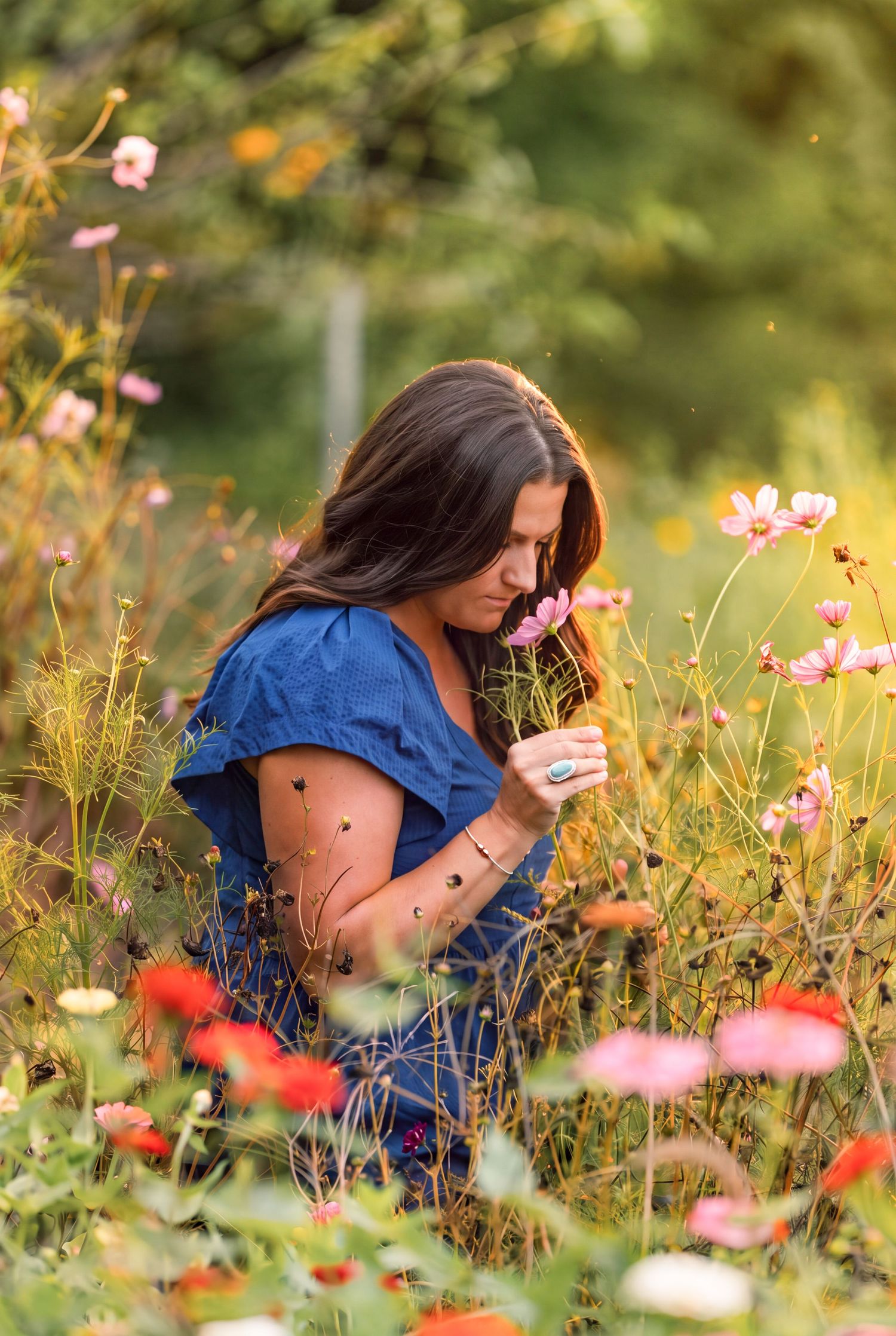 Brunette woman in blue dress kneeling among colorful wildflowers in a lush garden at dusk.