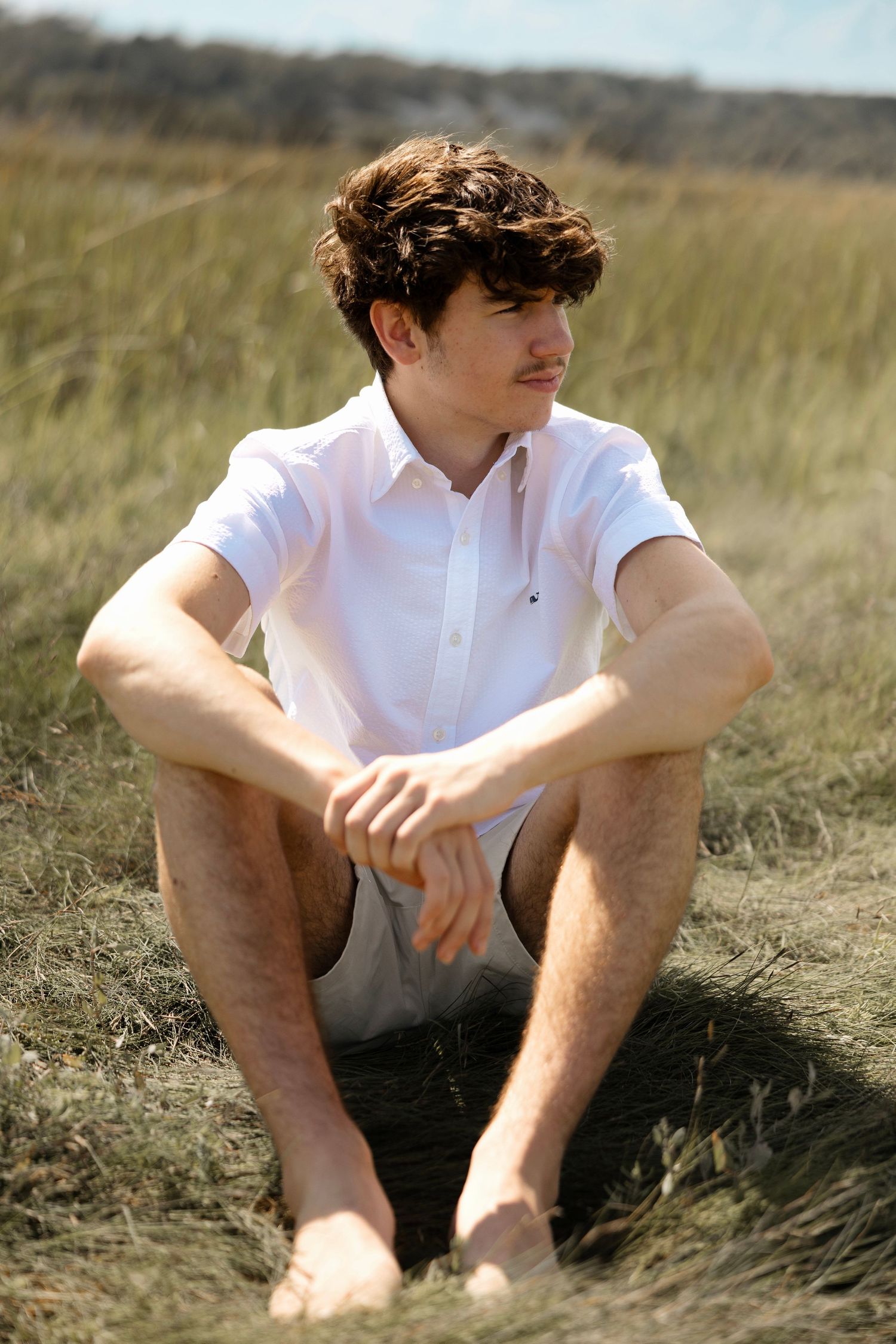 Young man in white shirt sitting barefoot in a grassy field on a sunny day.