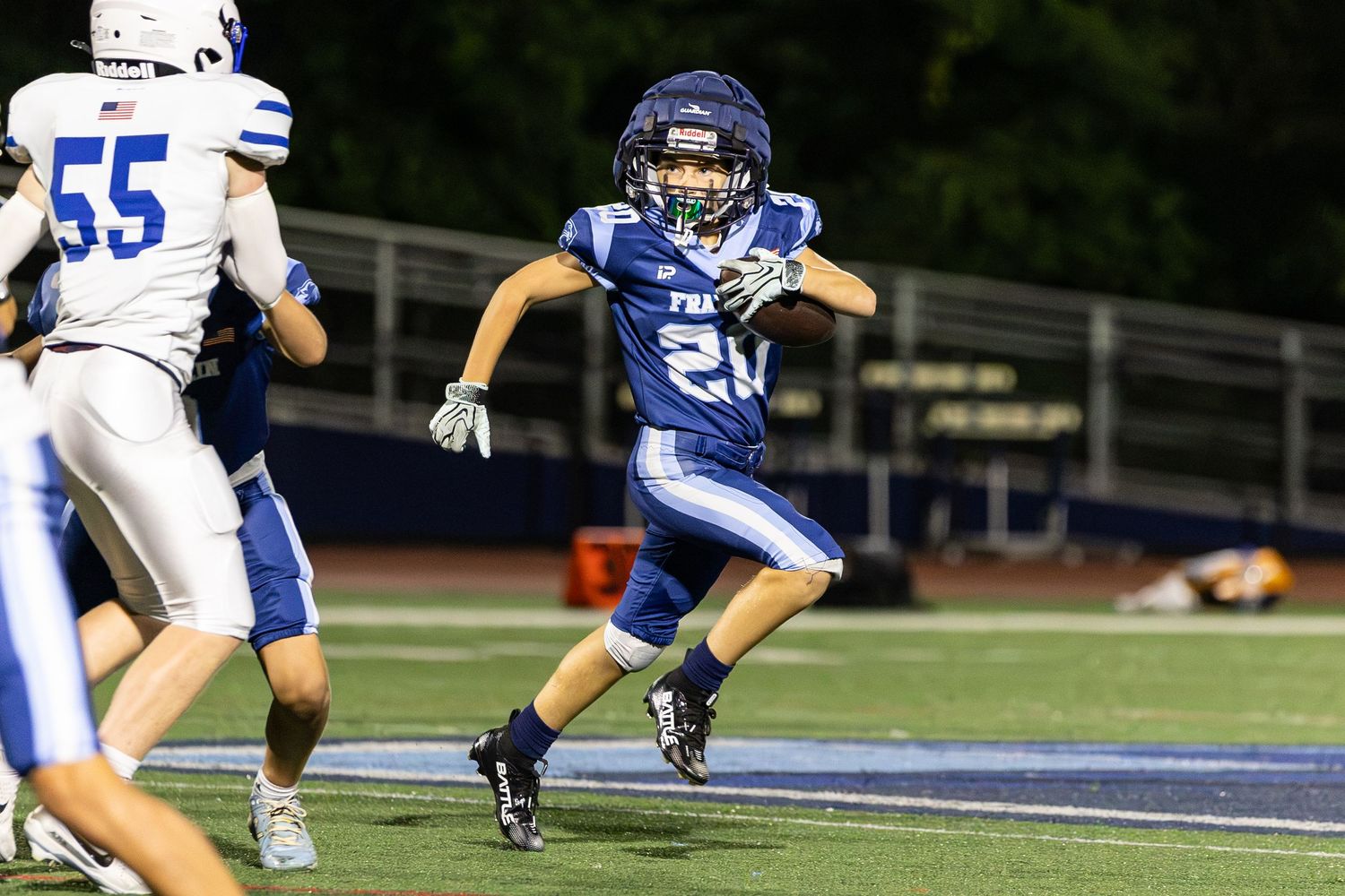 Youth football player in blue uniform number 21 runs downfield during a night game under bright stadium lights.