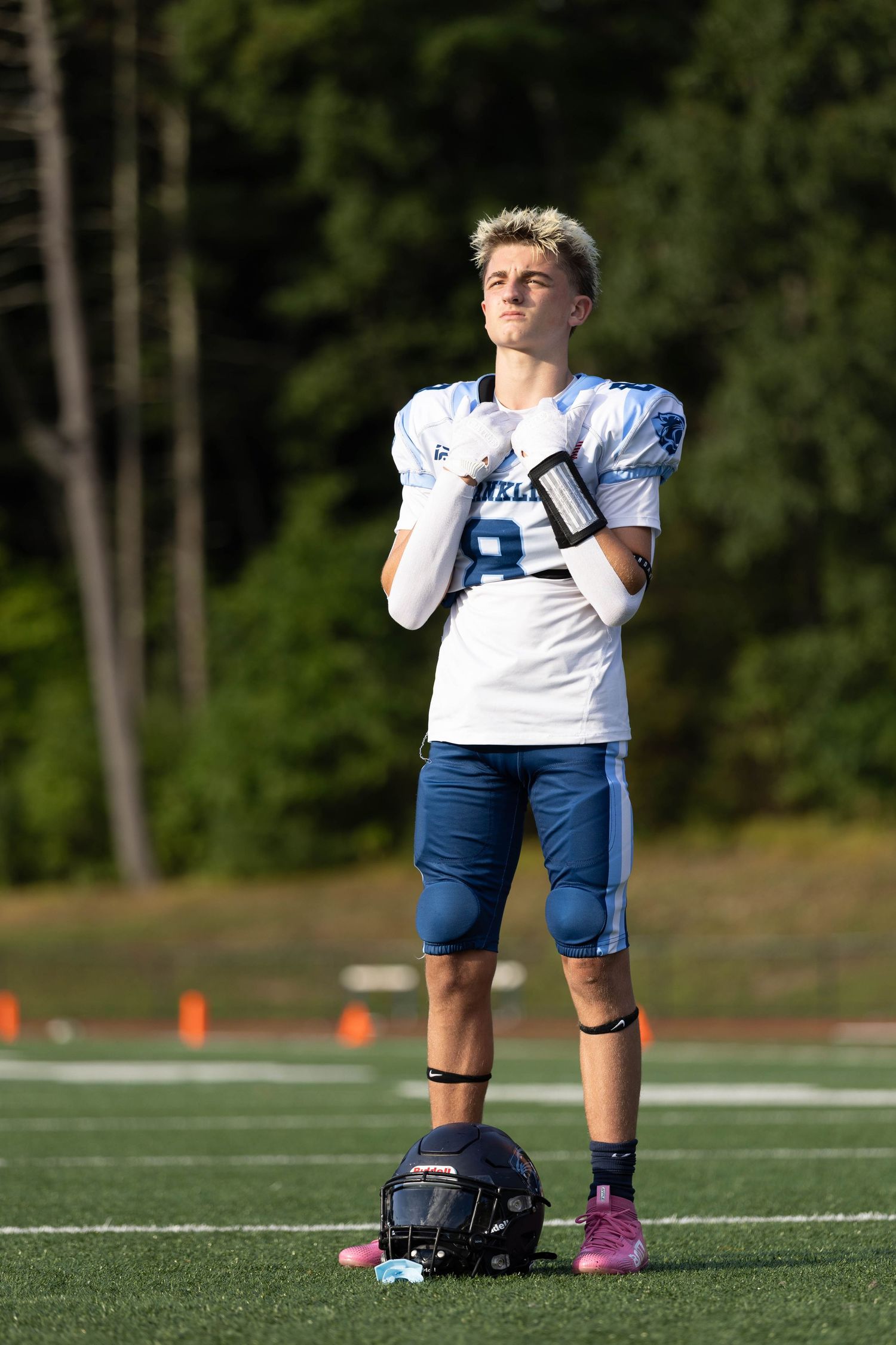 Young football player standing on field looking up, helmet at feet, sunny day.