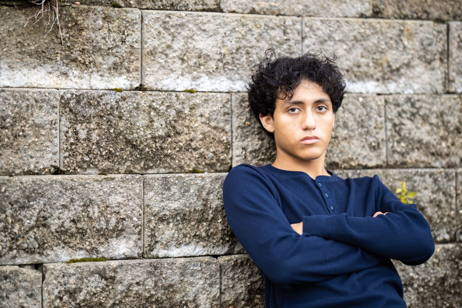 A young teen with curly hair leans against a stone wall with arms crossed, wearing a navy blue henley shirt.