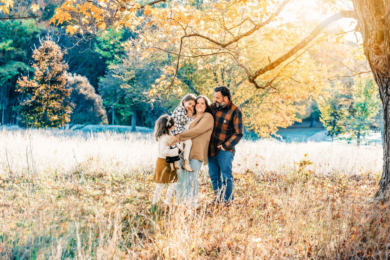 Family of four in warm autumn outfits embracing in a golden field beneath a tree with bright orange fall leaves.