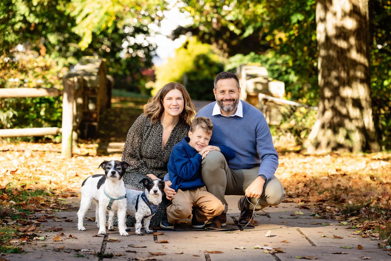 Smiling family of three crouching on a park path in autumn with two small black and white dogs, surrounded by fall foliage.