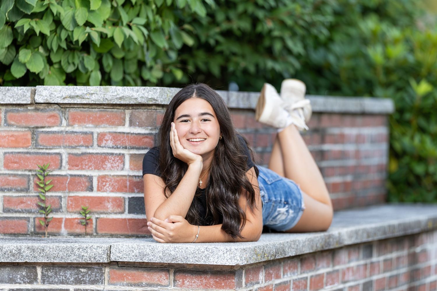 Smiling teenage girl in black top and denim shorts lying on a brick wall surrounded by green foliage.