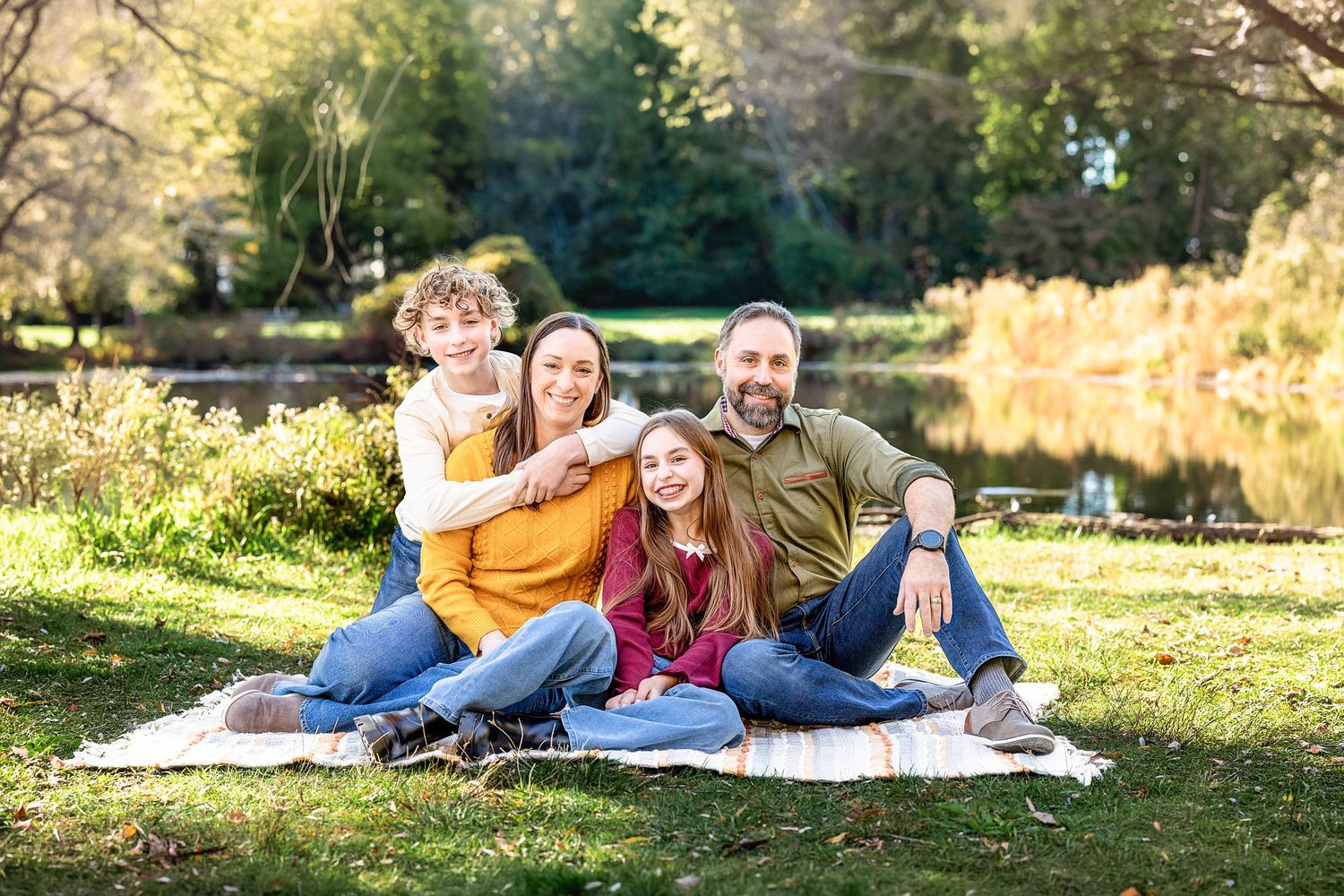 Smiling family of four sits together on a picnic blanket in a sunny park beside a calm pond surrounded by autumn trees.
