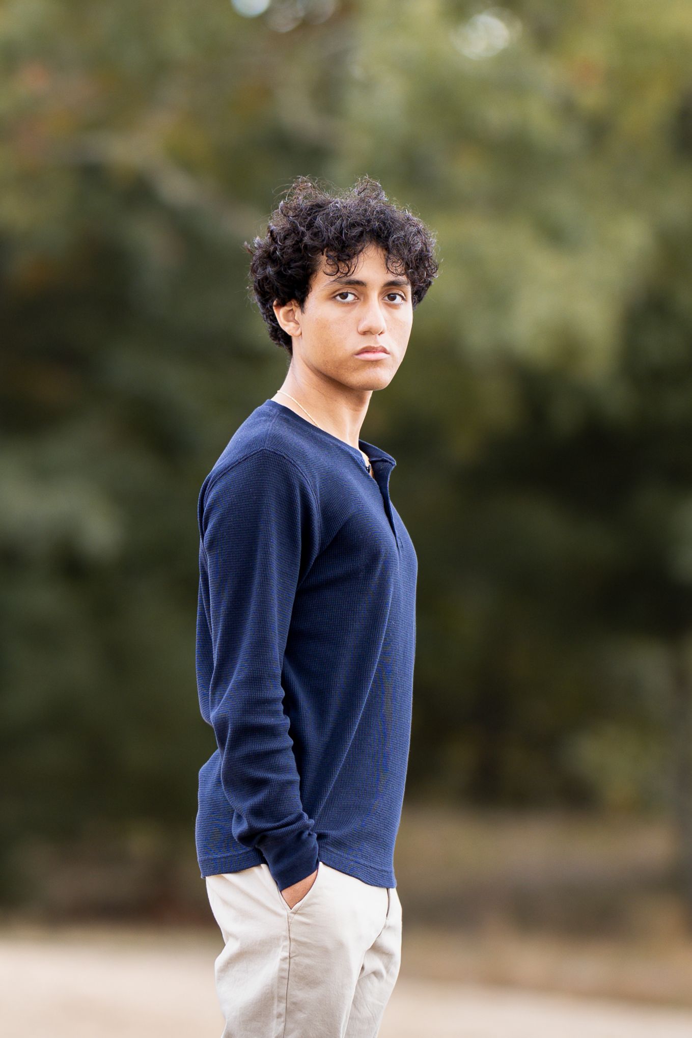 A young teen boy with curly hair wearing a navy blue sweater and khaki pants stands outdoors near trees.