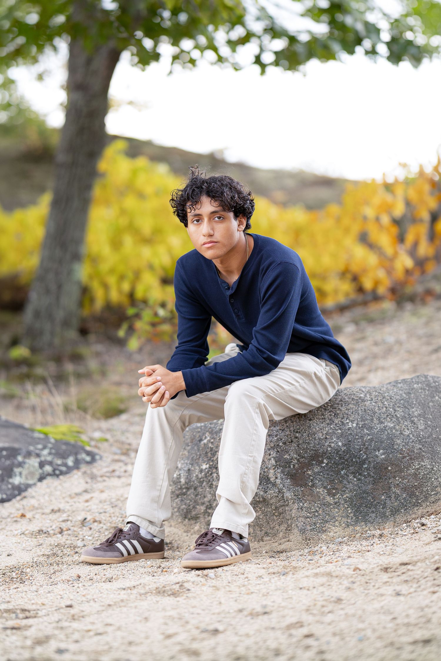 Teen boy in navy sweater and white pants sits on a rock outdoors with autumn foliage in the background.