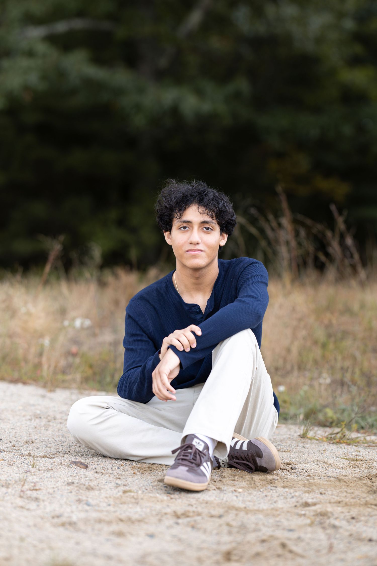 Young man with curly hair sits cross-legged on a dirt path wearing a navy sweater and white pants outdoors.