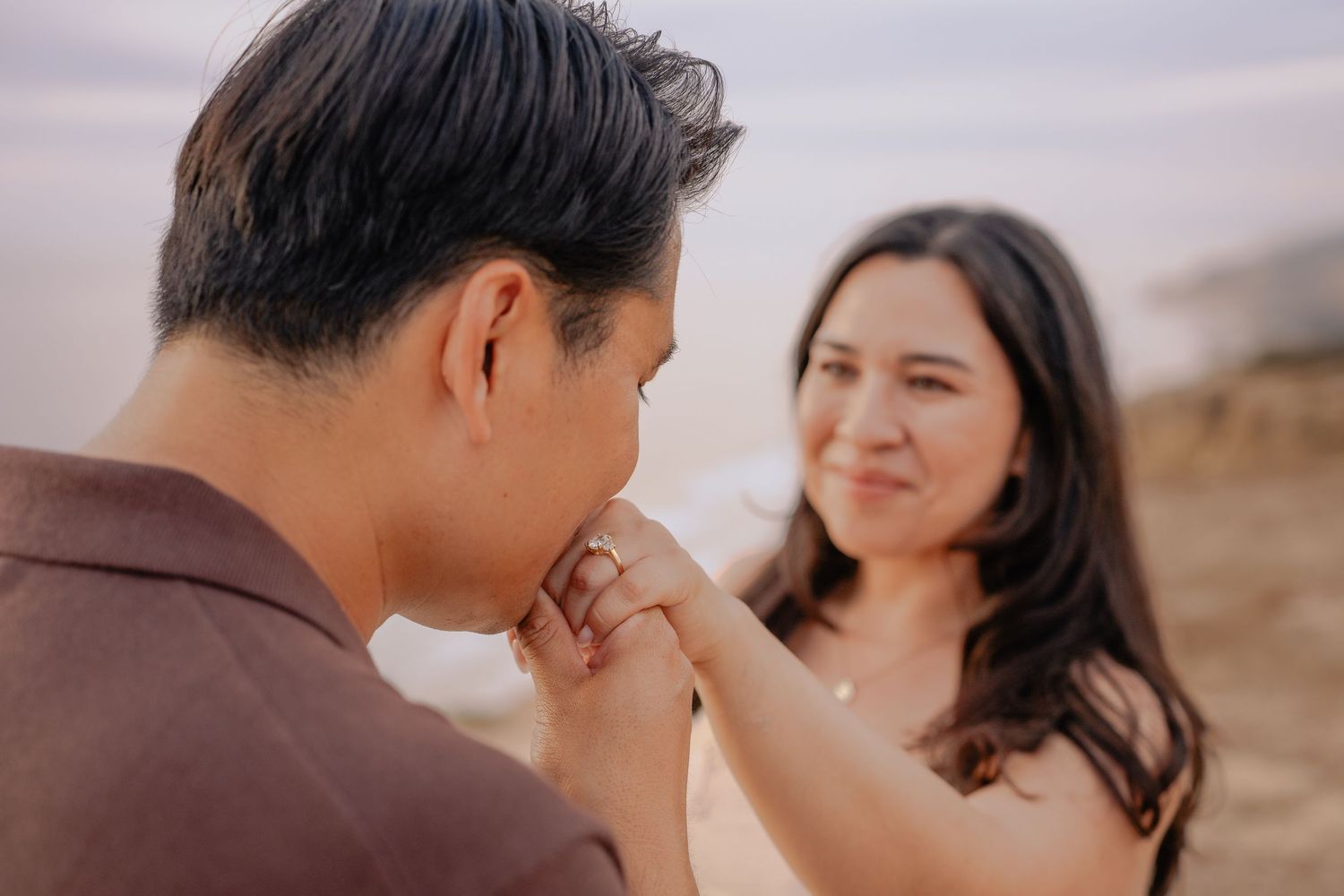 A man kisses a woman's hand on a beach at sunset during a romantic engagement photo session.