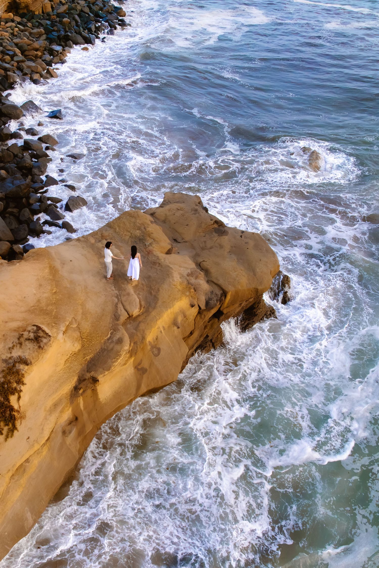 Aerial view of couple in white standing on rocky coastal cliff surrounded by crashing ocean waves. santa cruz cliffs san diego