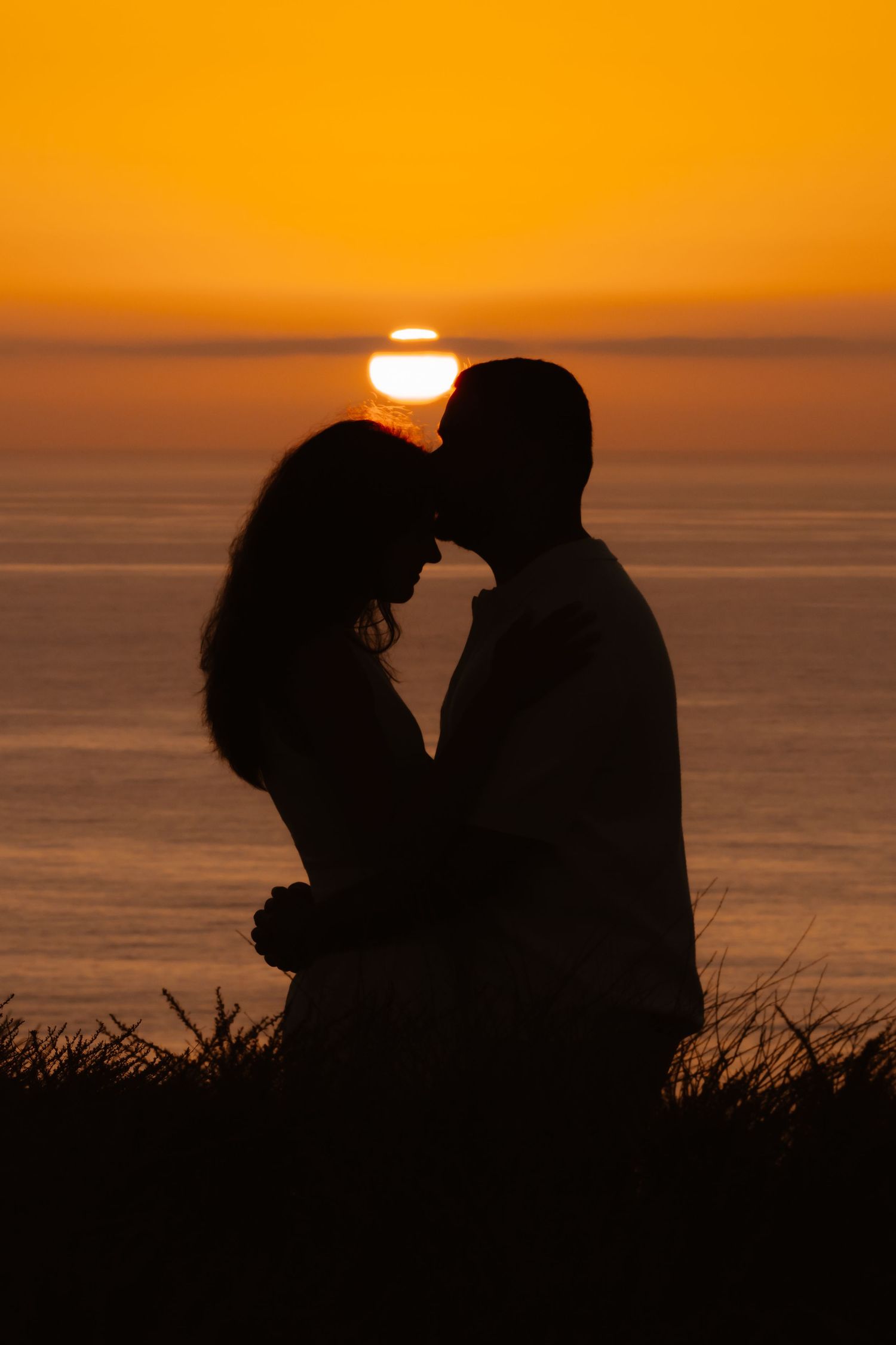 Romantic silhouette of couple about to kiss with vibrant orange sunset and ocean in background.