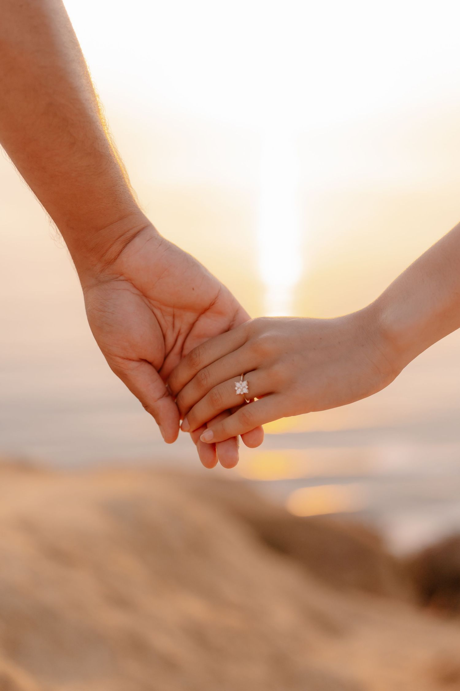 Couple holding hands at sunset, her sparkling diamond engagement ring visible against a glowing golden beach backdrop.