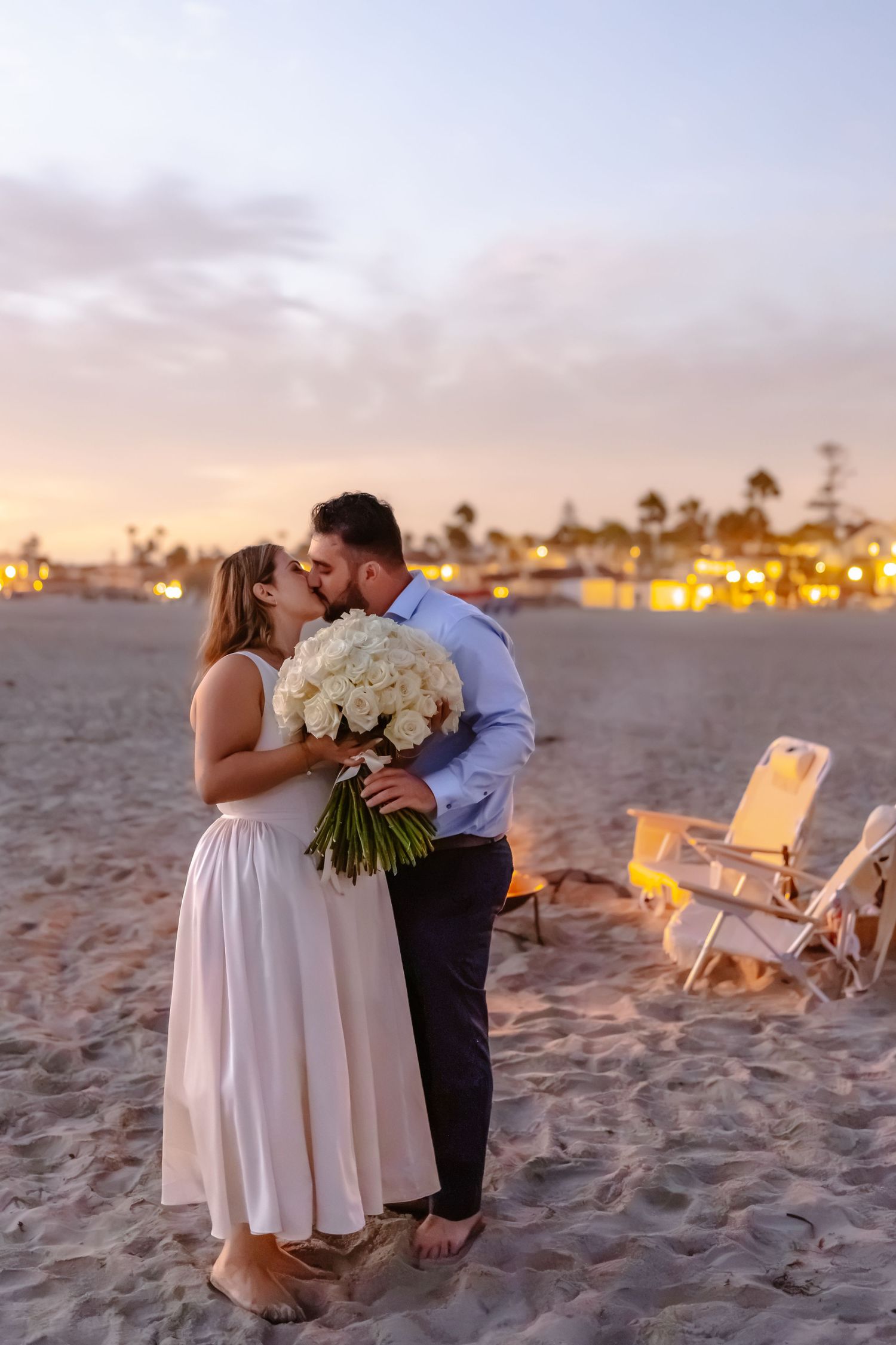 Engaged couple kissing on beach at sunset holding white rose bouquet. Coronado beach