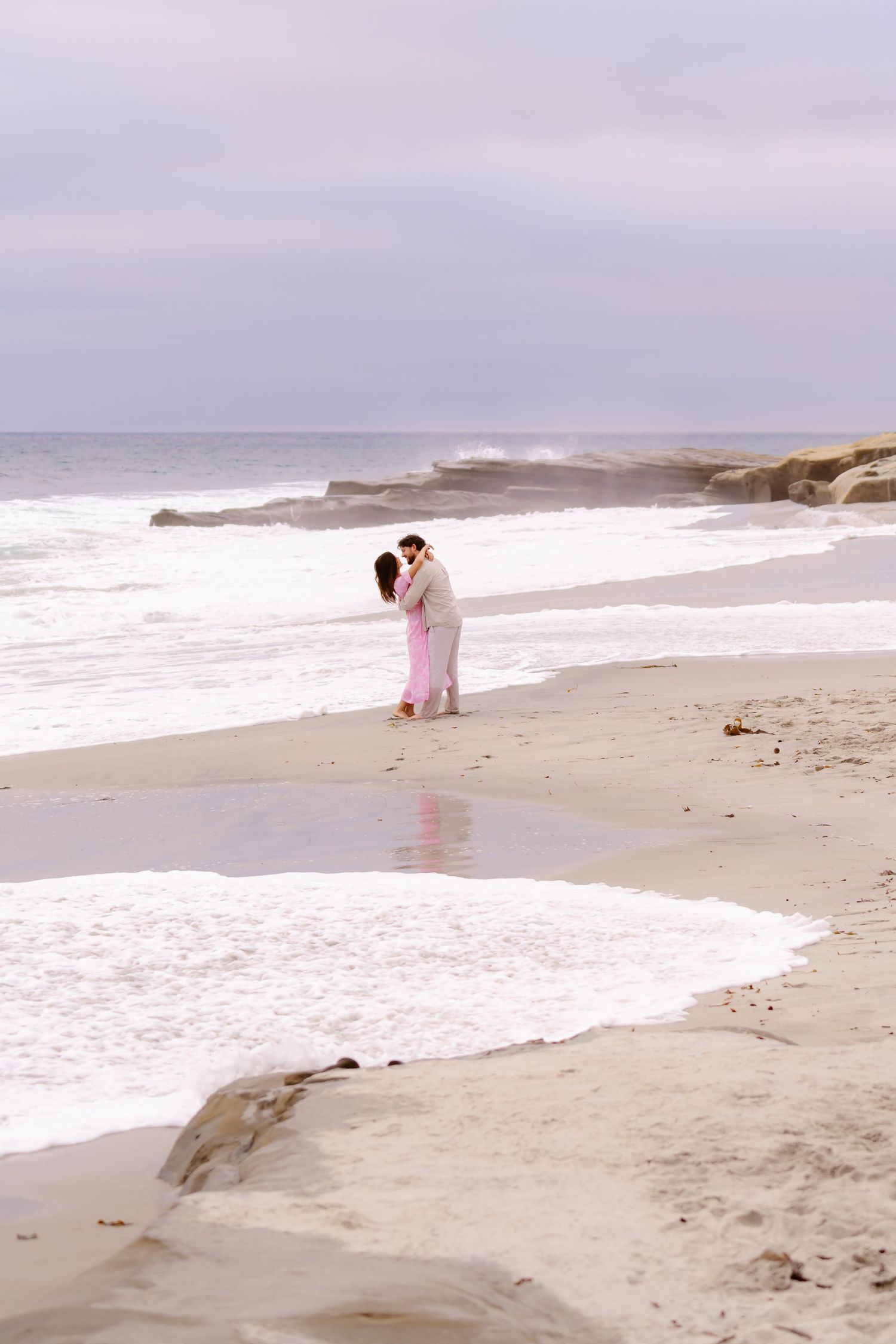 Couple kissing on wet sandy beach with ocean waves and rocky shore behind them.