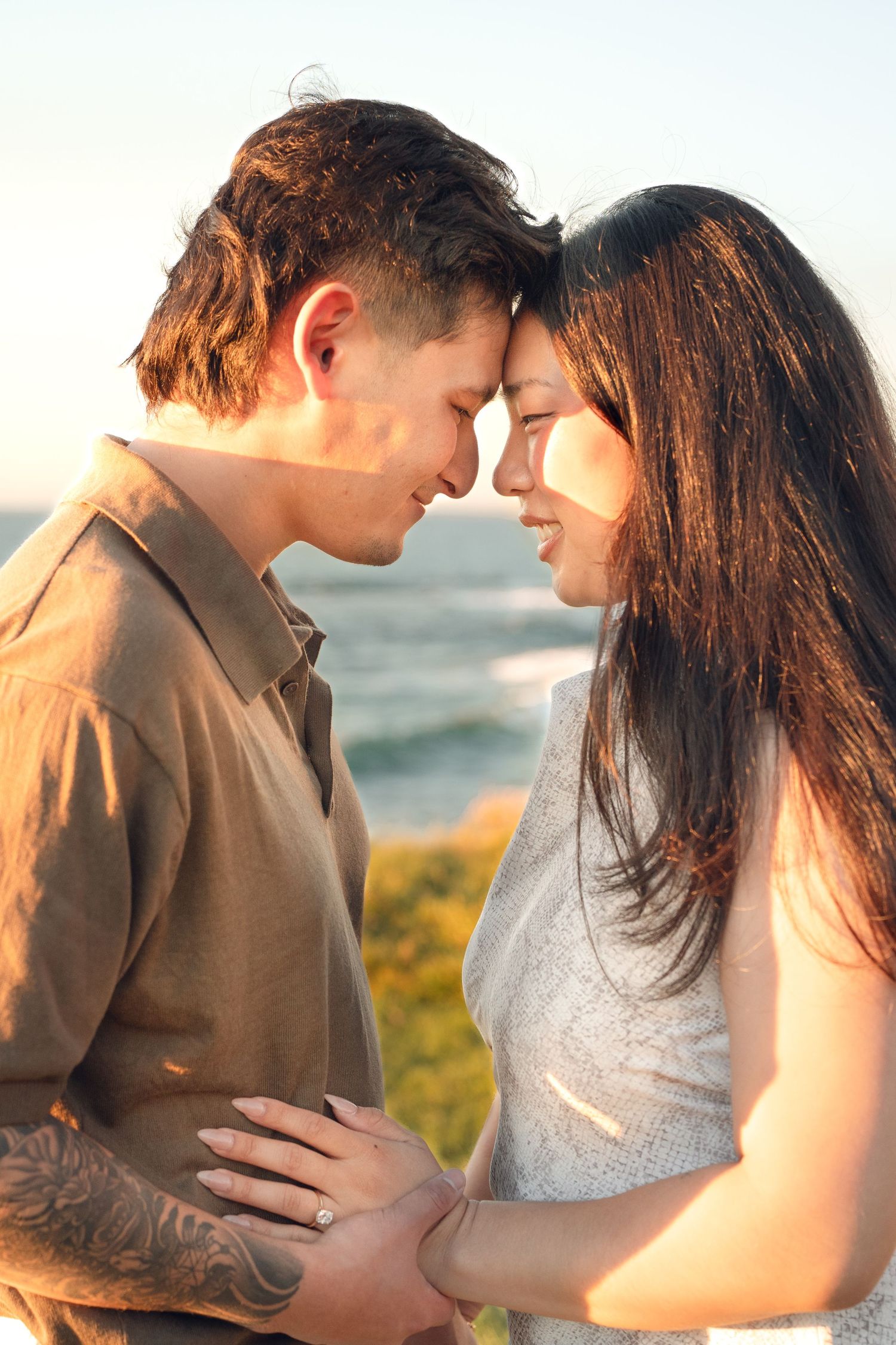 Couple touching foreheads and smiling at sunset beside the ocean, woman wearing a ring and man with a forearm tattoo.