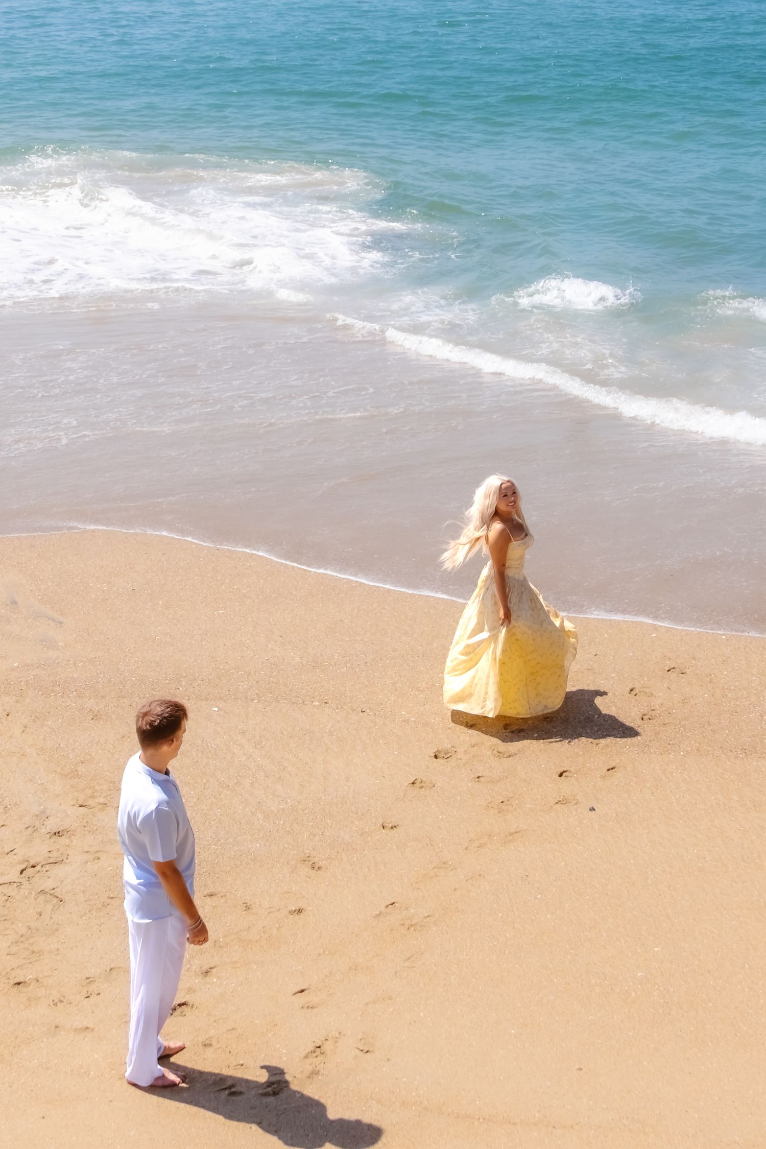 Woman in flowing yellow dress near ocean waves while man in white watches from sandy beach on a sunny day.