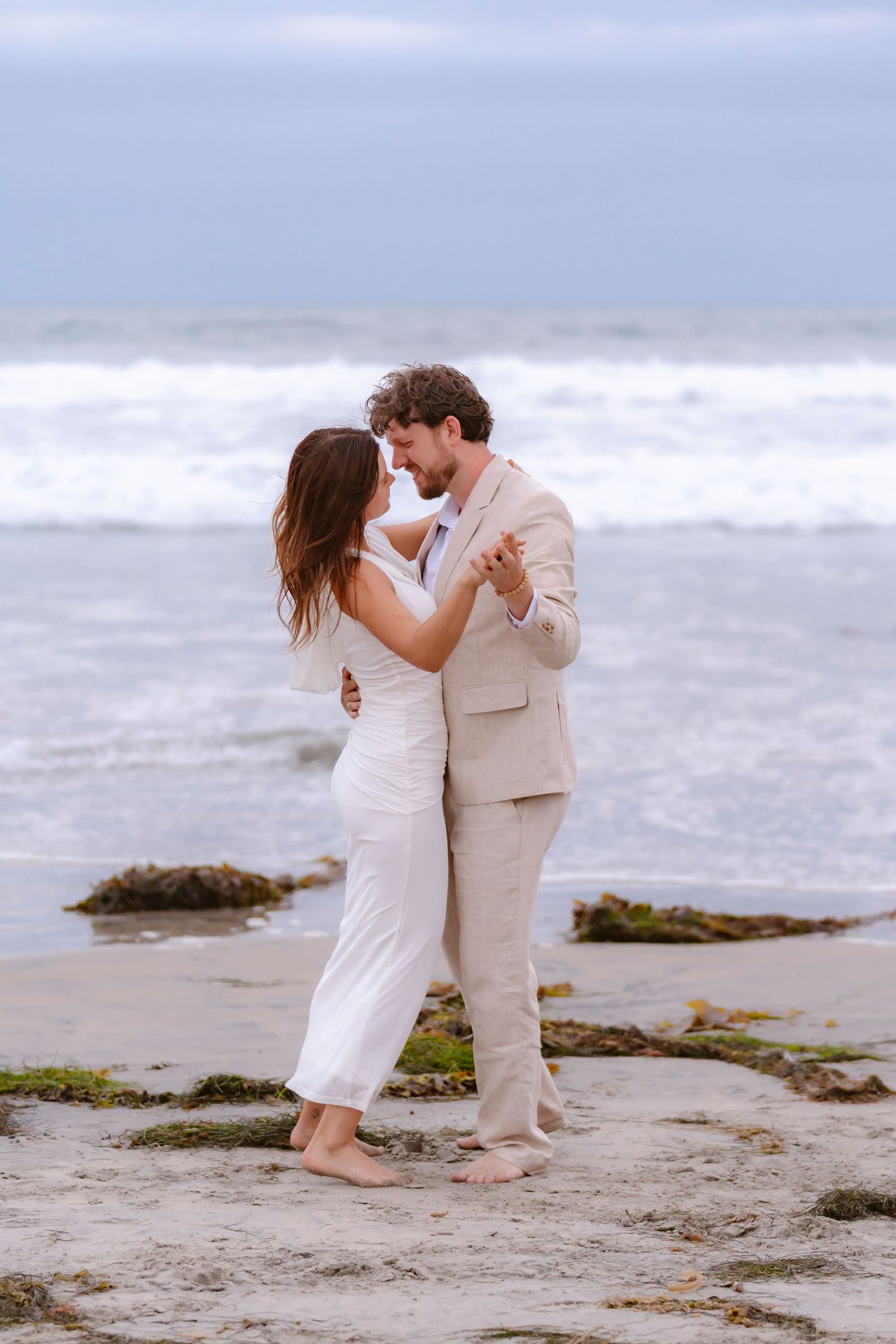 Couple slow dancing barefoot on sandy beach near ocean, woman in white outfit and man in beige suit.