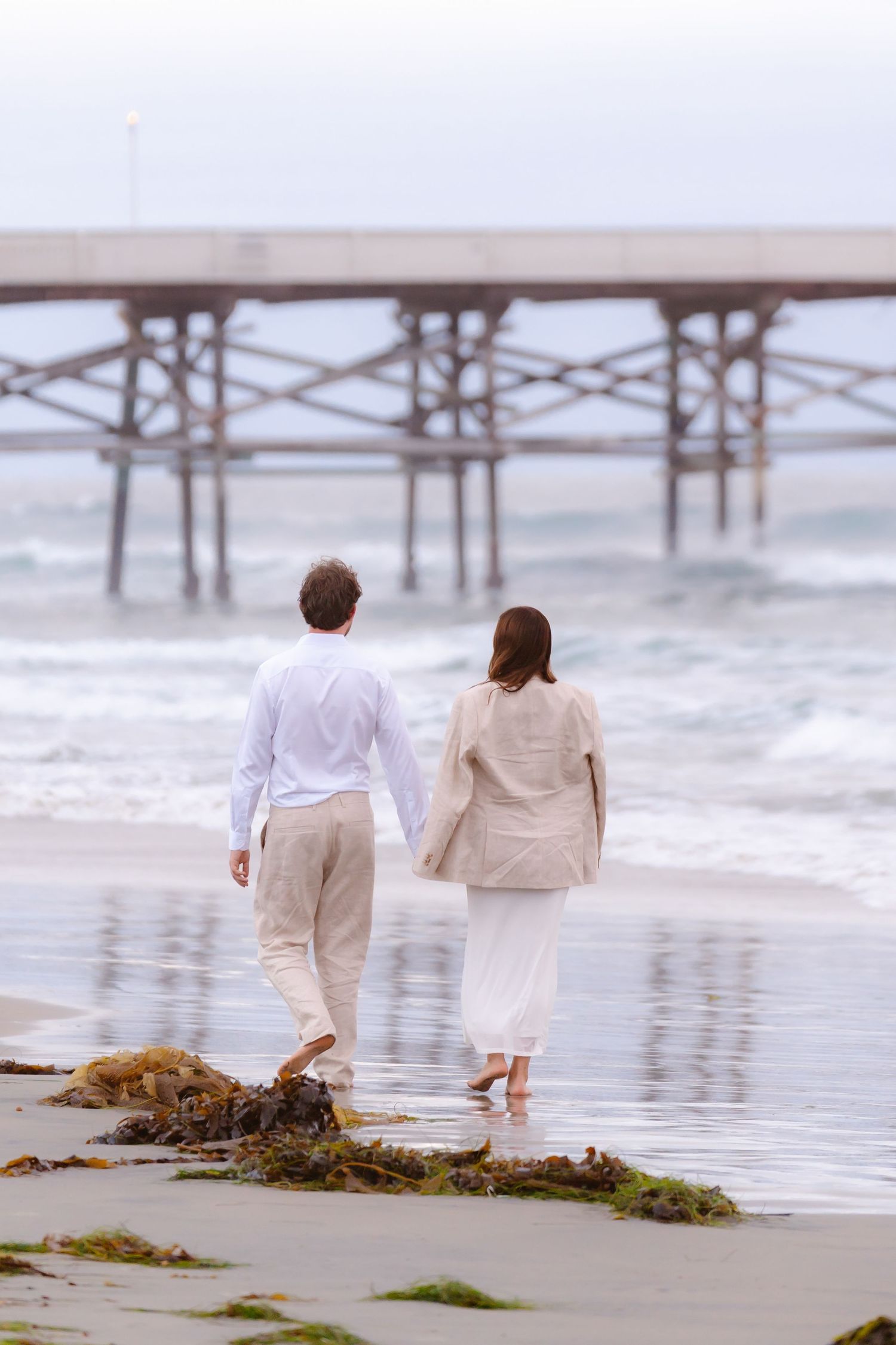 Couple walking hand in hand barefoot along a beach toward a pier, dressed in white and beige outfits.