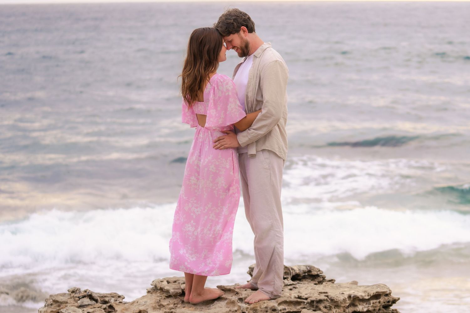 Romantic couple embracing on rocky coastal cliff, woman in pink floral dress and man in beige suit by the sea.