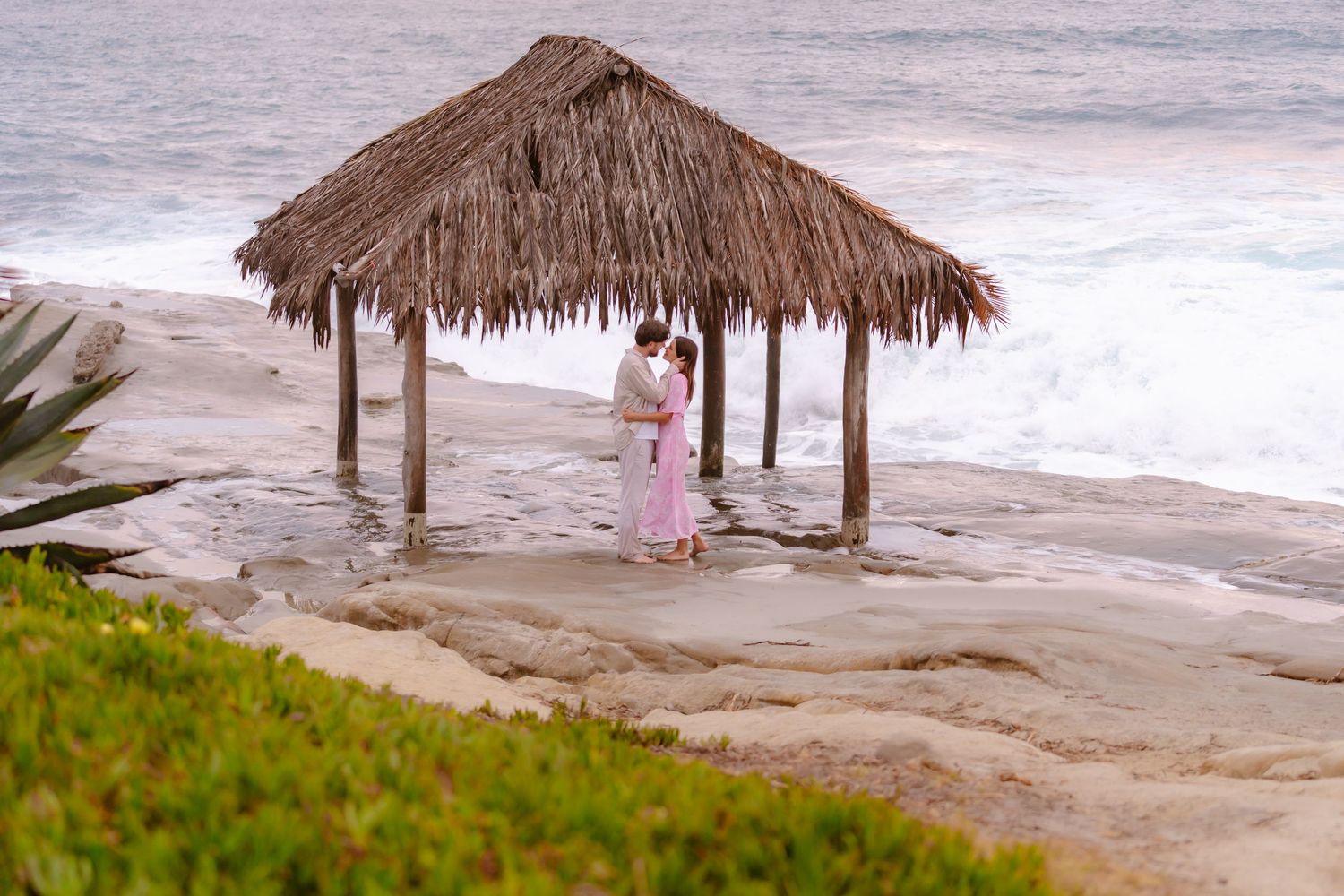 Romantic couple kissing under a thatched palapa hut on rocky shoreline with ocean waves in the background.