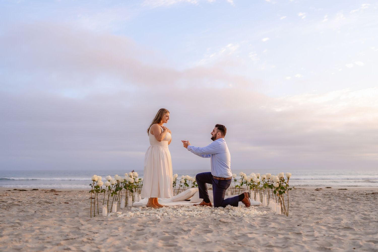 Man proposes to woman on a romantic beach setup with white flowers at sunset.