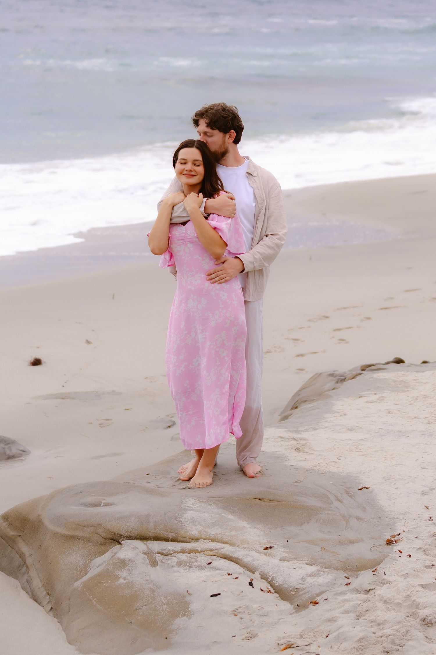 Man kissing woman on cheek while embracing on rocky beach, woman in pink dress.