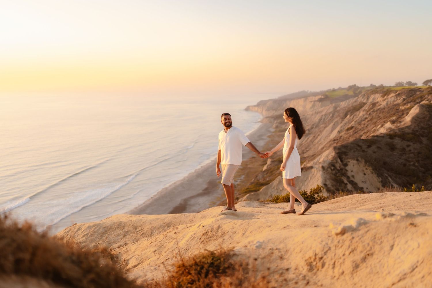 Couple in white holding hands walking along dramatic coastal cliffs with ocean view at sunset. Torrey Pines