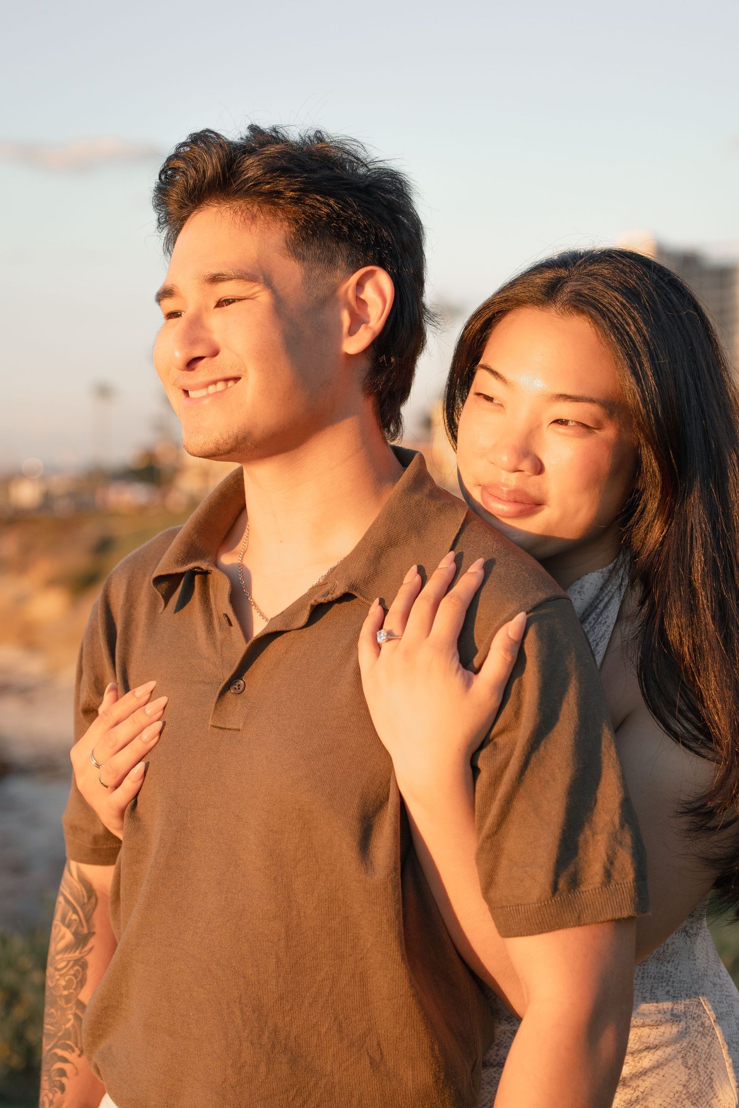 Smiling couple embracing outdoors at golden hour, the woman wearing a ring and resting her hand on the man's shoulder.