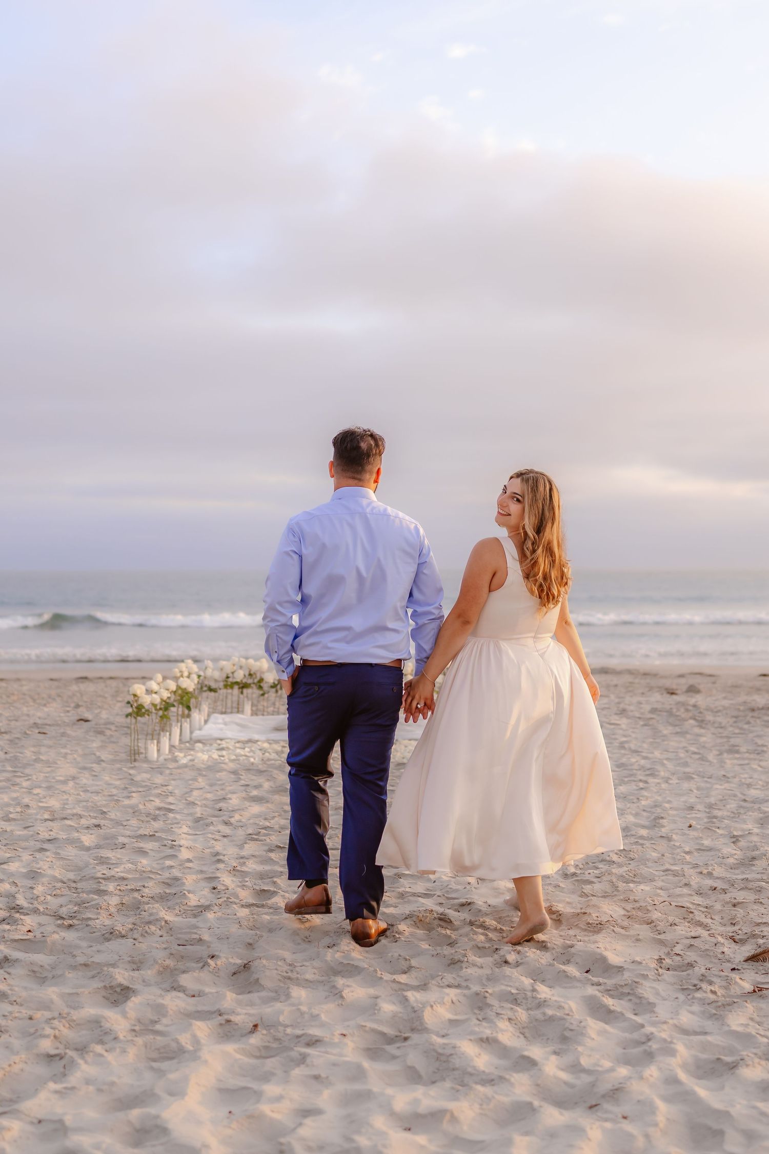 Couple holding hands walking toward Coronado beach proposal flower arrangement at sunset.