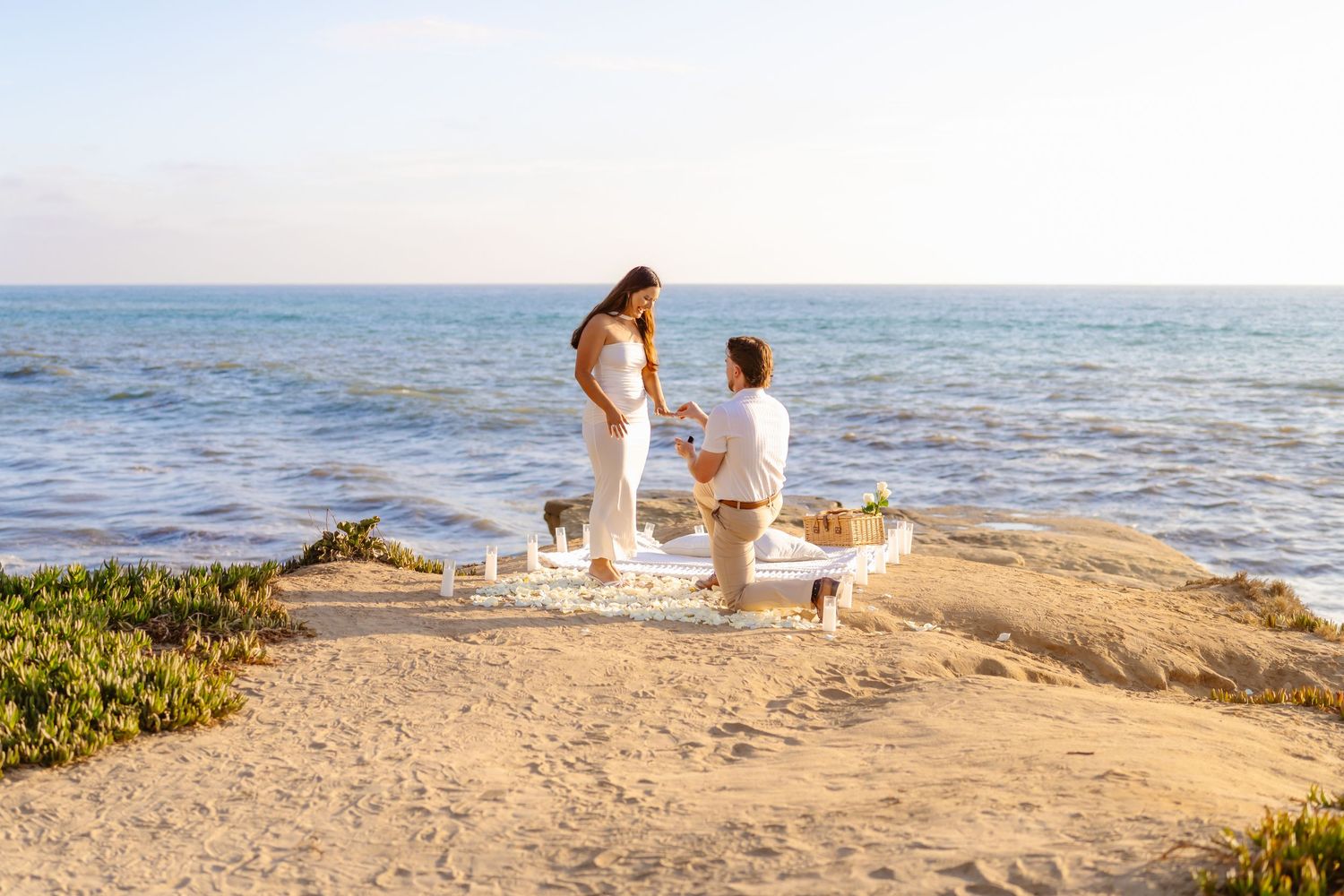 Man proposing on one knee to woman at romantic beach picnic setup by ocean Santa Cruz Cliffs