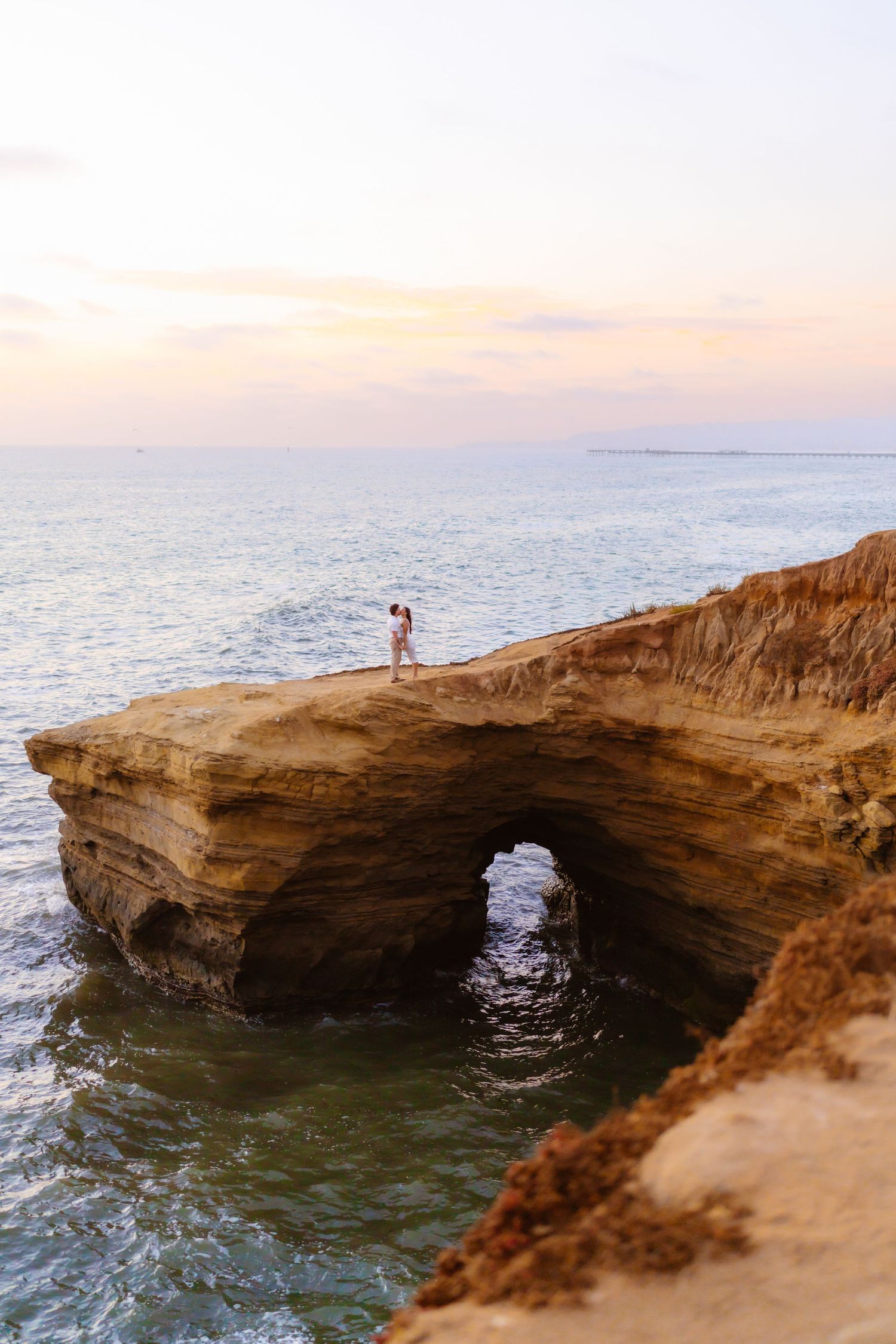 Couple standing on a natural rock arch over the ocean at sunset in golden coastal cliffs.