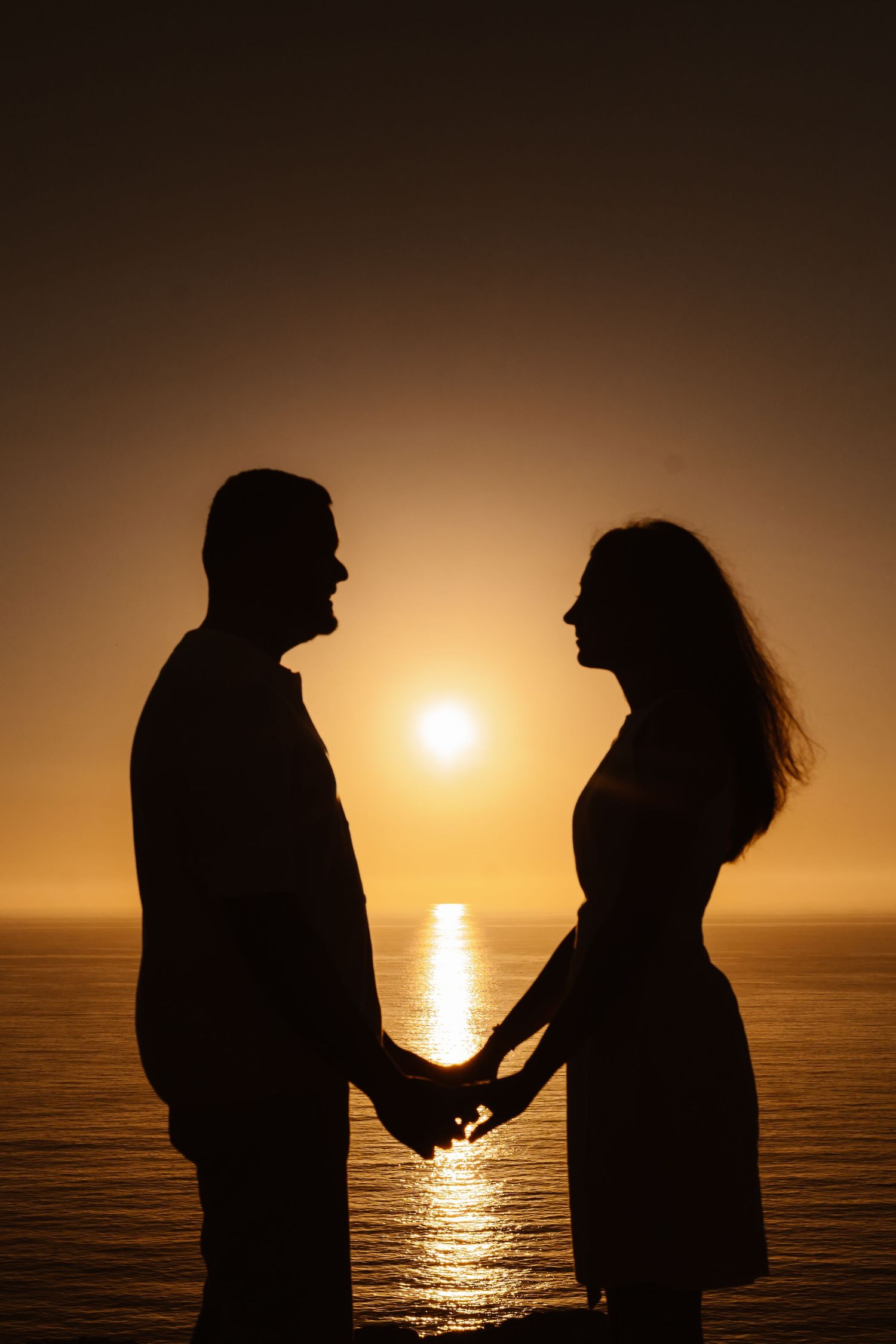 Romantic silhouette of couple holding hands facing each other with golden sunset reflecting on ocean.