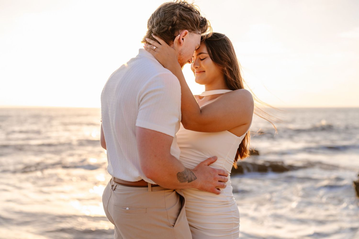 Romantic couple embracing forehead to forehead at sunset with ocean waves behind them.