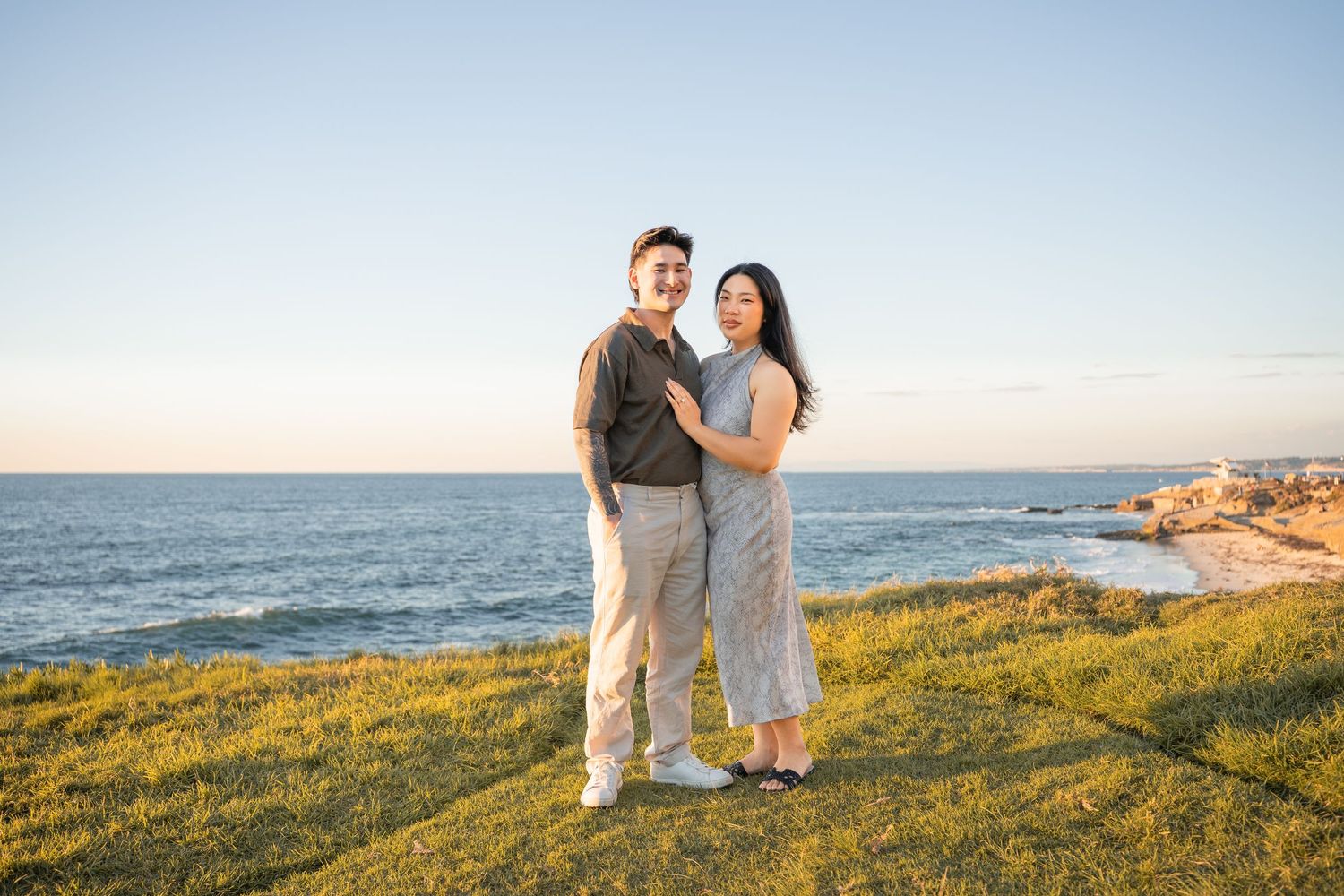Smiling couple embracing on a grassy coastal cliff at sunset with the ocean and rocky shoreline stretching behind them.