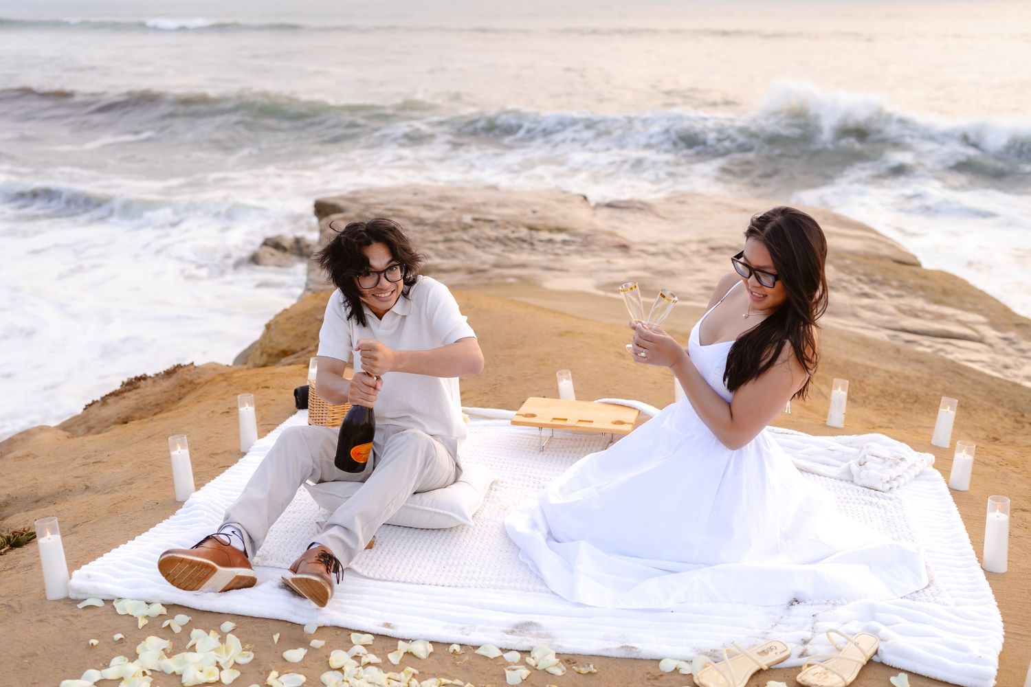 Couple relaxing on white blanket with candles and champagne on rocky oceanside at sunset. santa cruz cliffs san diego
