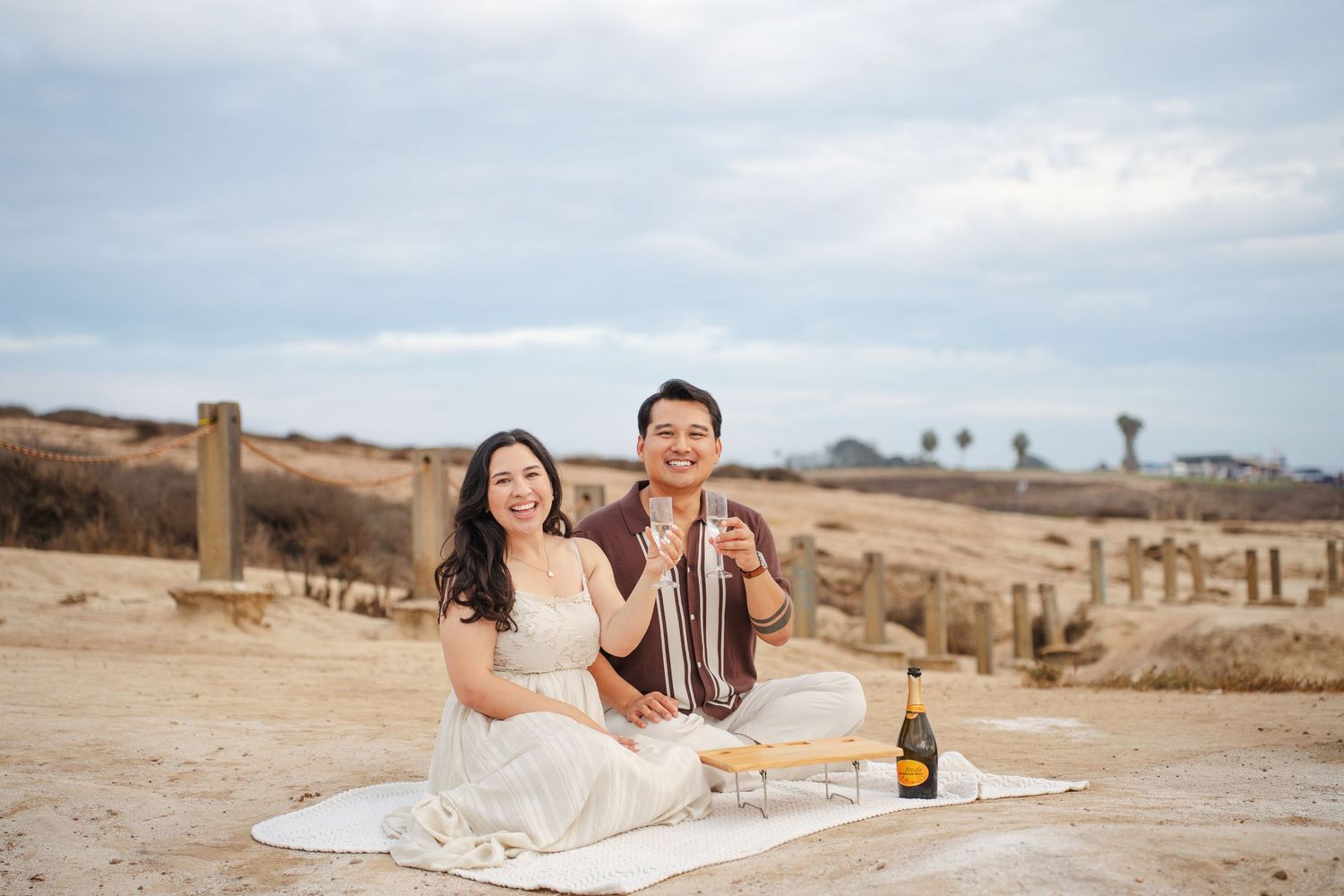 Couple smiling and toasting with champagne on a sandy beach blanket picnic under a cloudy sky.