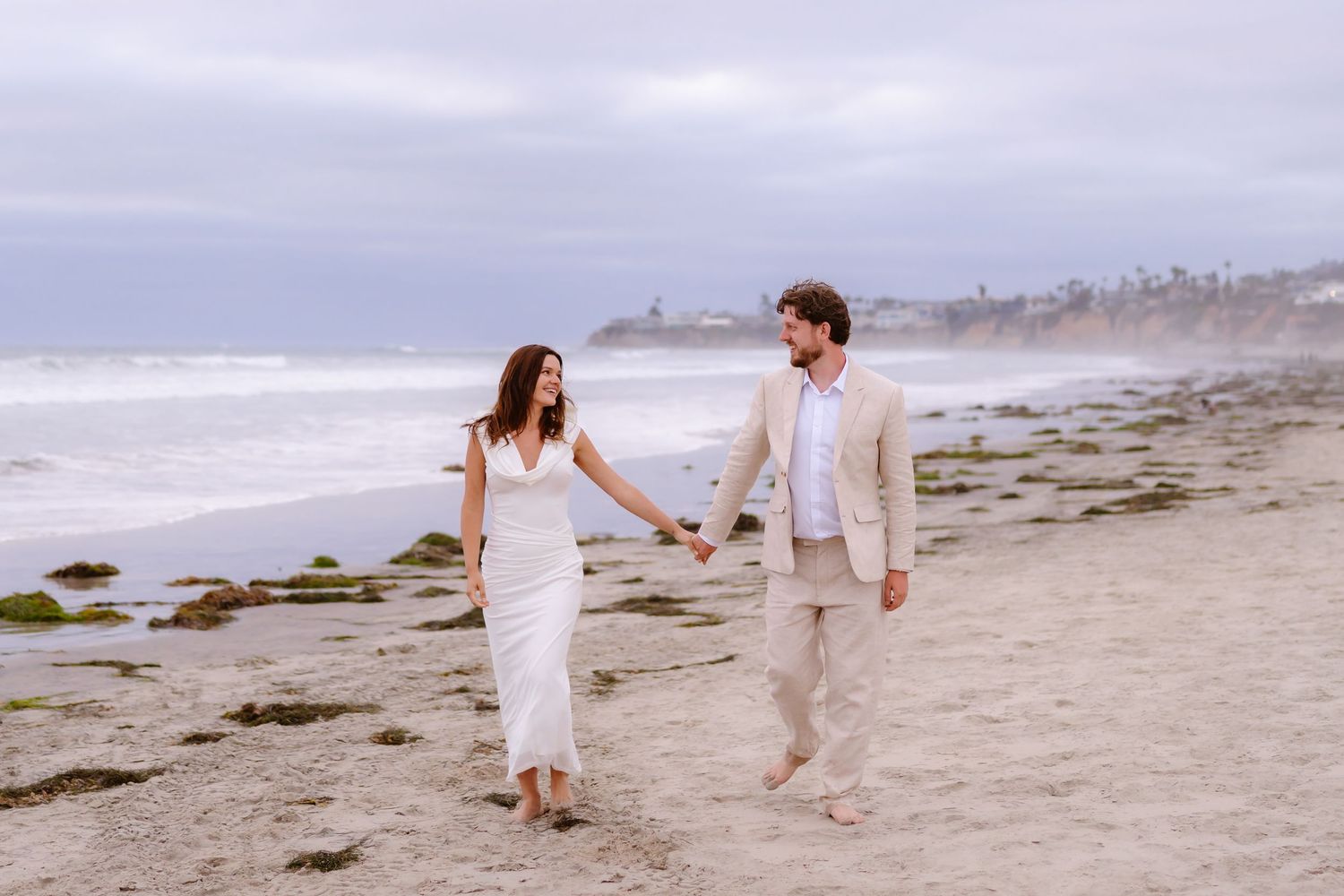 Smiling couple holding hands and walking on a sandy beach, woman in white dress and man in beige suit by the ocean.