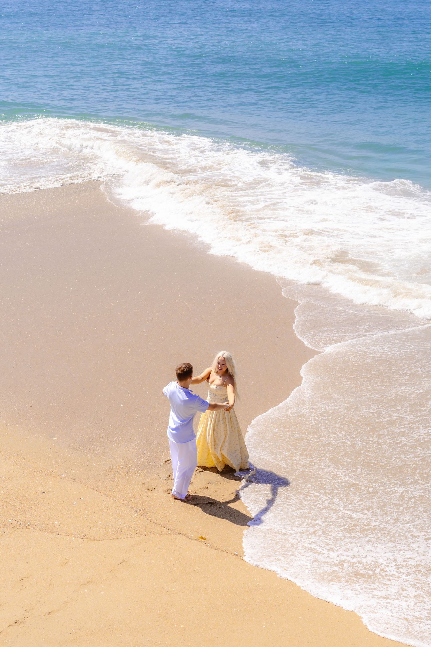 Aerial view of couple holding hands on sandy beach with turquoise ocean waves.