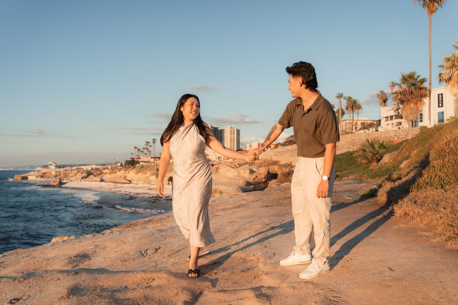 Couple holding hands and walking along a sunny rocky coastal bluff with ocean views, palm trees, and seaside buildings in the background.