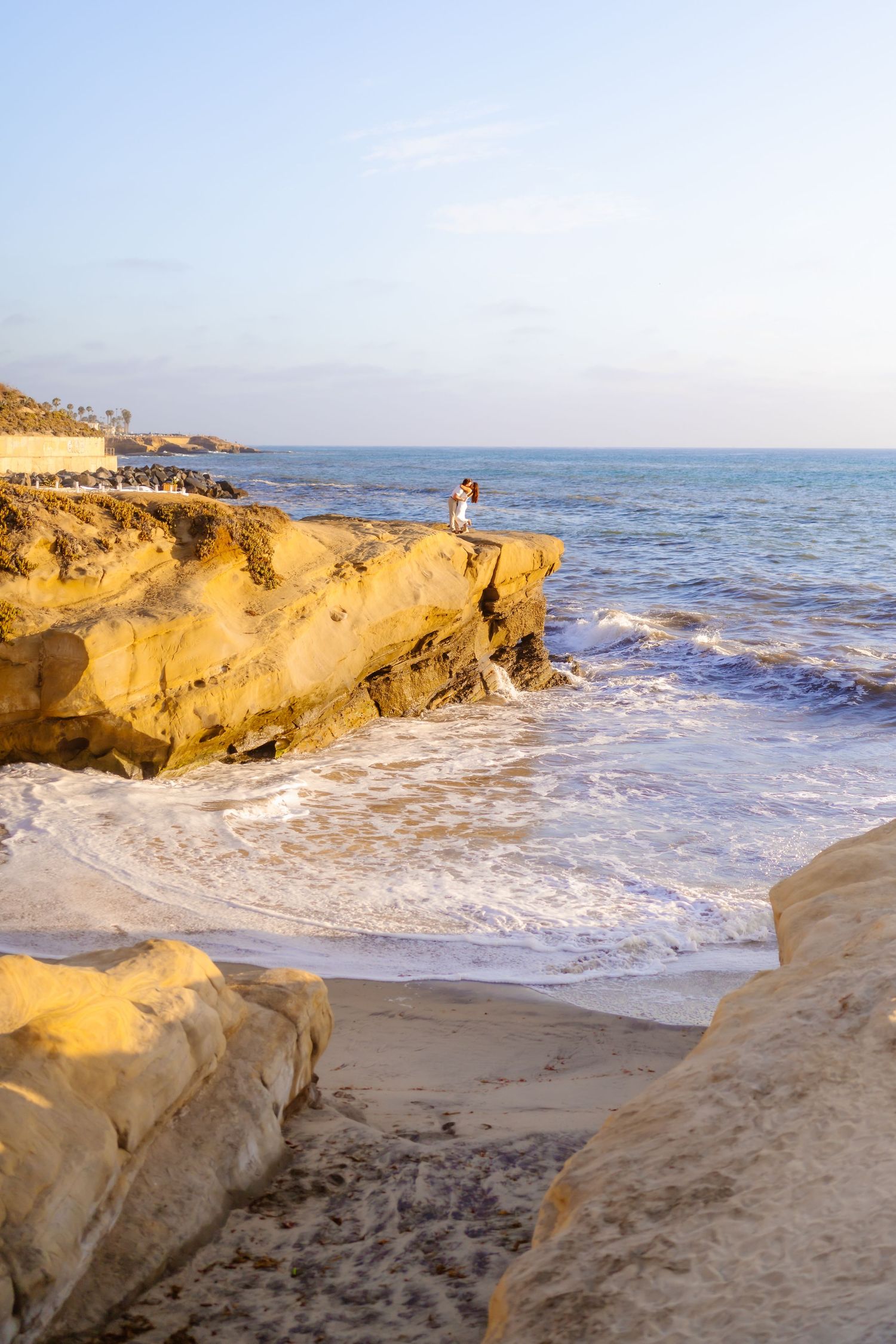Couple standing on rocky coastal cliffs embracing with ocean waves crashing below. Santa Cruz Cliffs