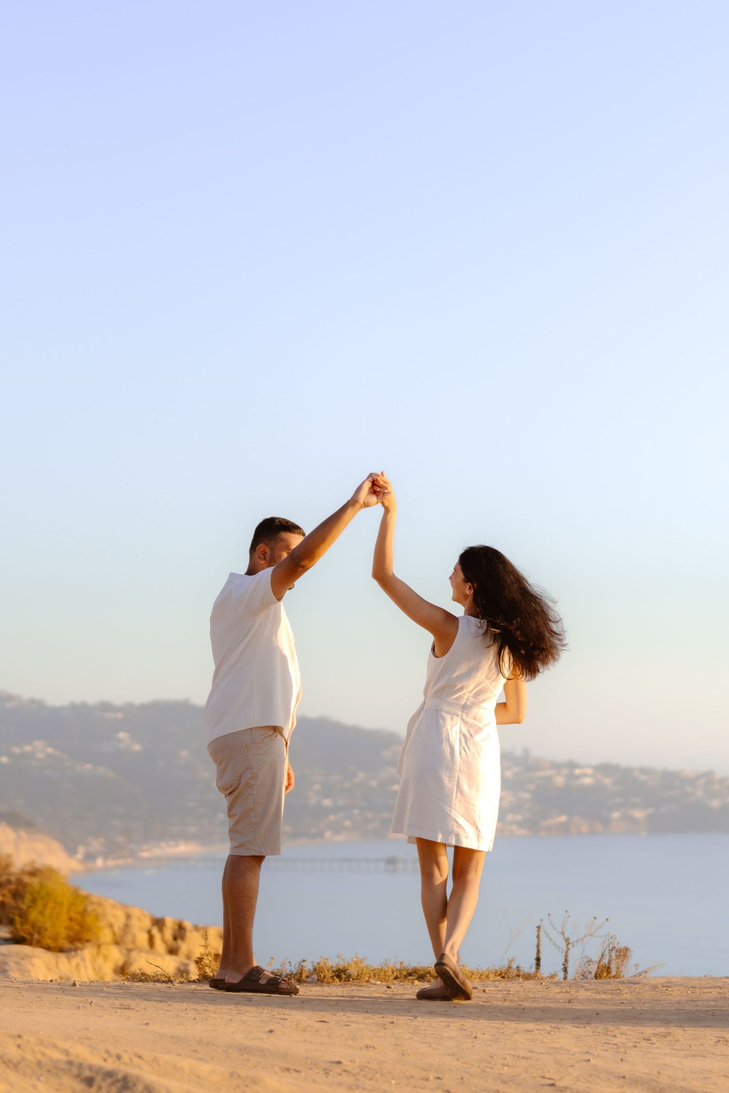 Couple in white dancing joyfully on sandy hilltop with blue ocean and clear sky in background.