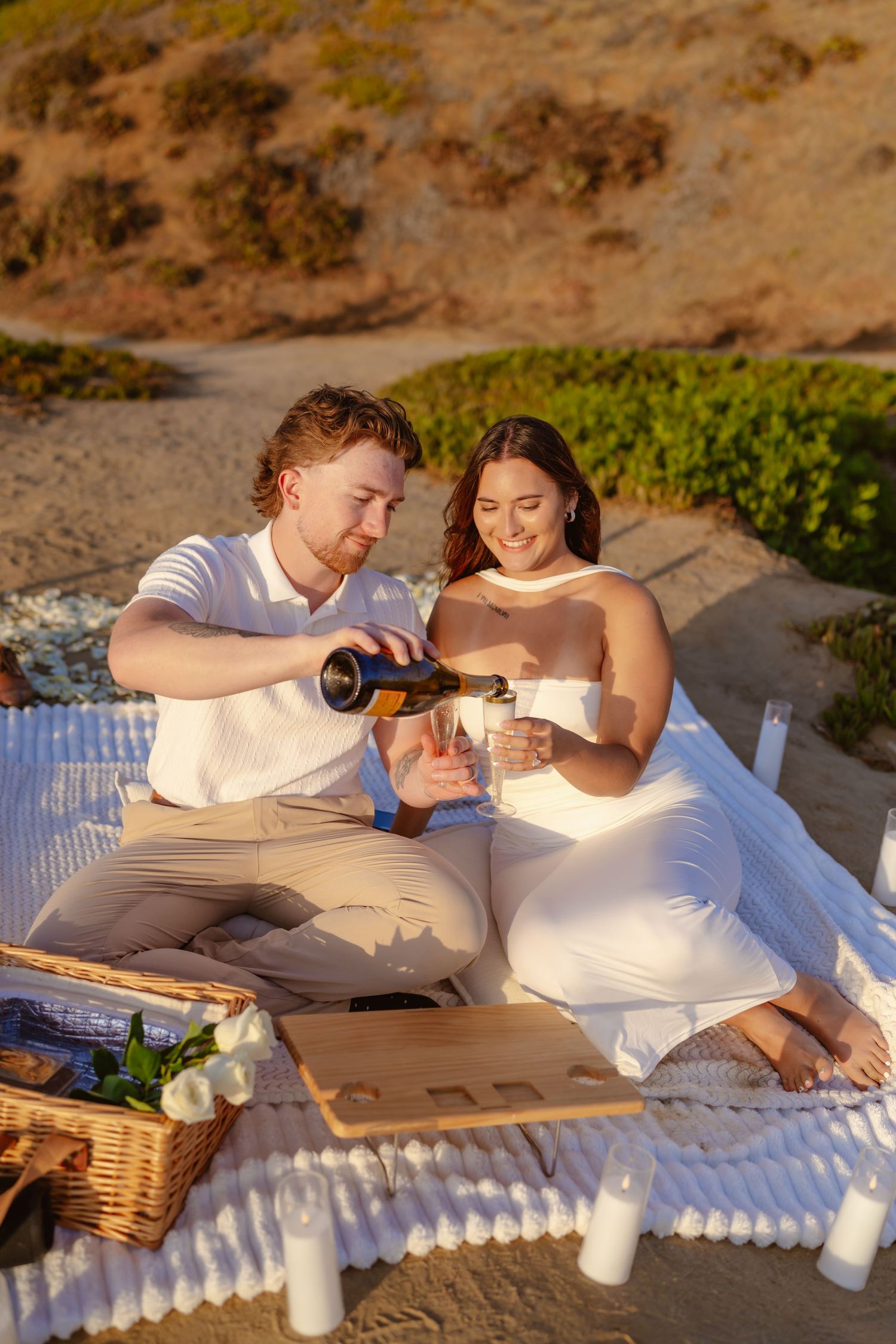 Couple sharing champagne on a picnic blanket at beach during golden sunset hour. Santa Cruz Cliffs