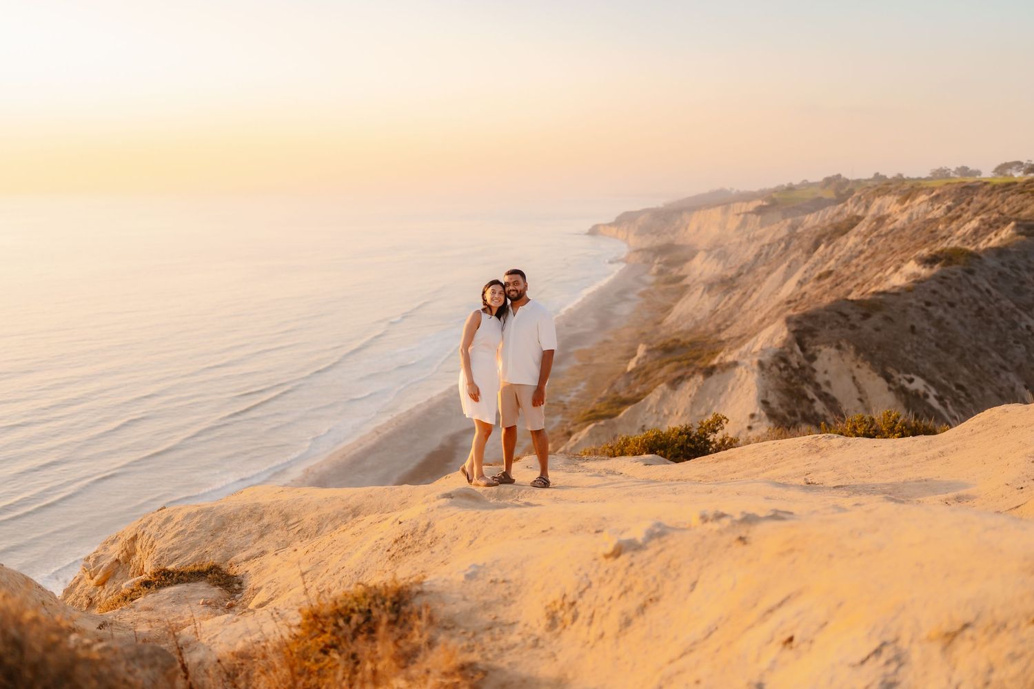 Happy couple in matching white outfits embracing on sandy cliffs with ocean coastline at sunset. Torrey Pines