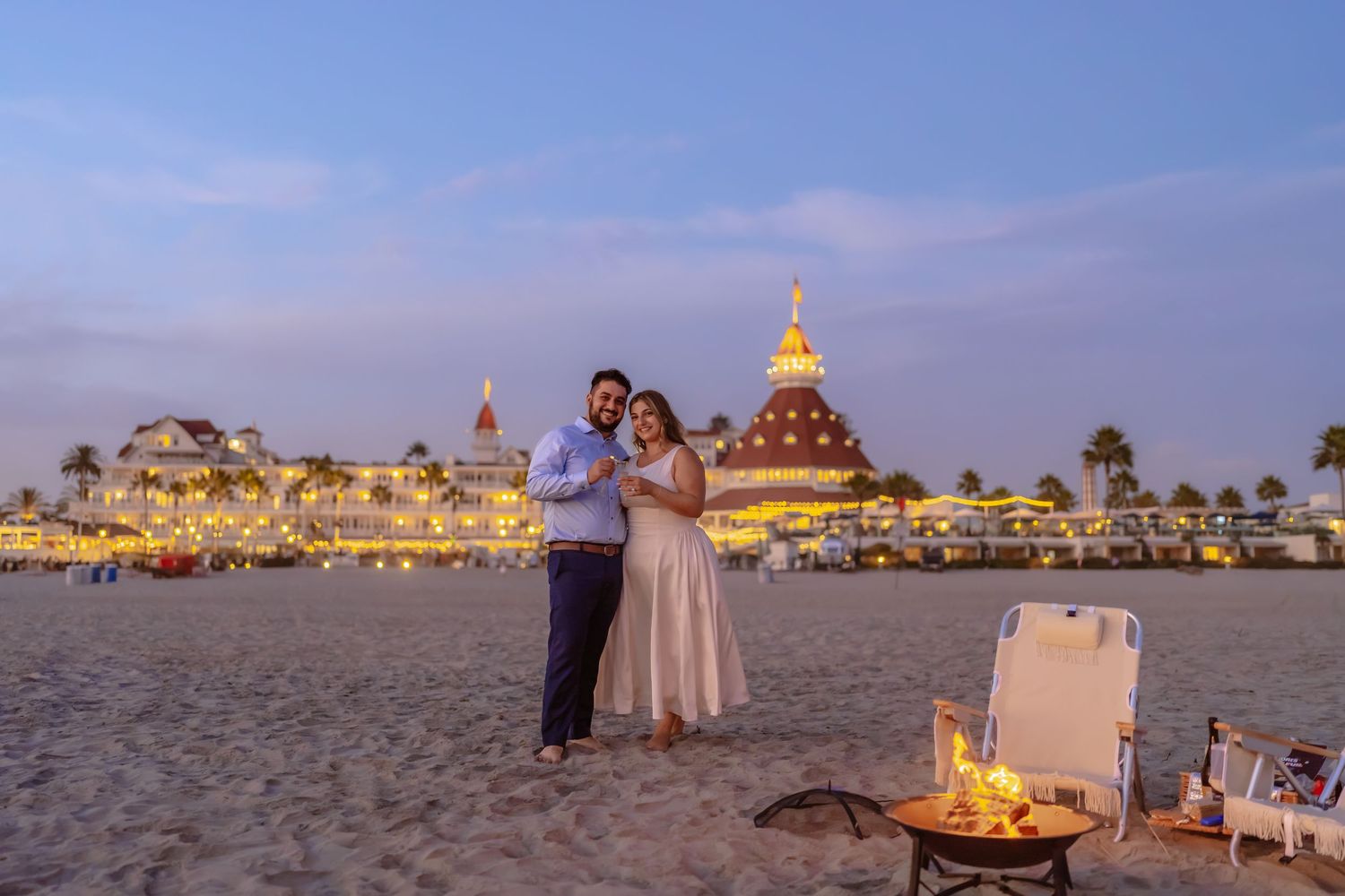 Couple embracing on beach at dusk with Hotel del Coronado lit up behind them.