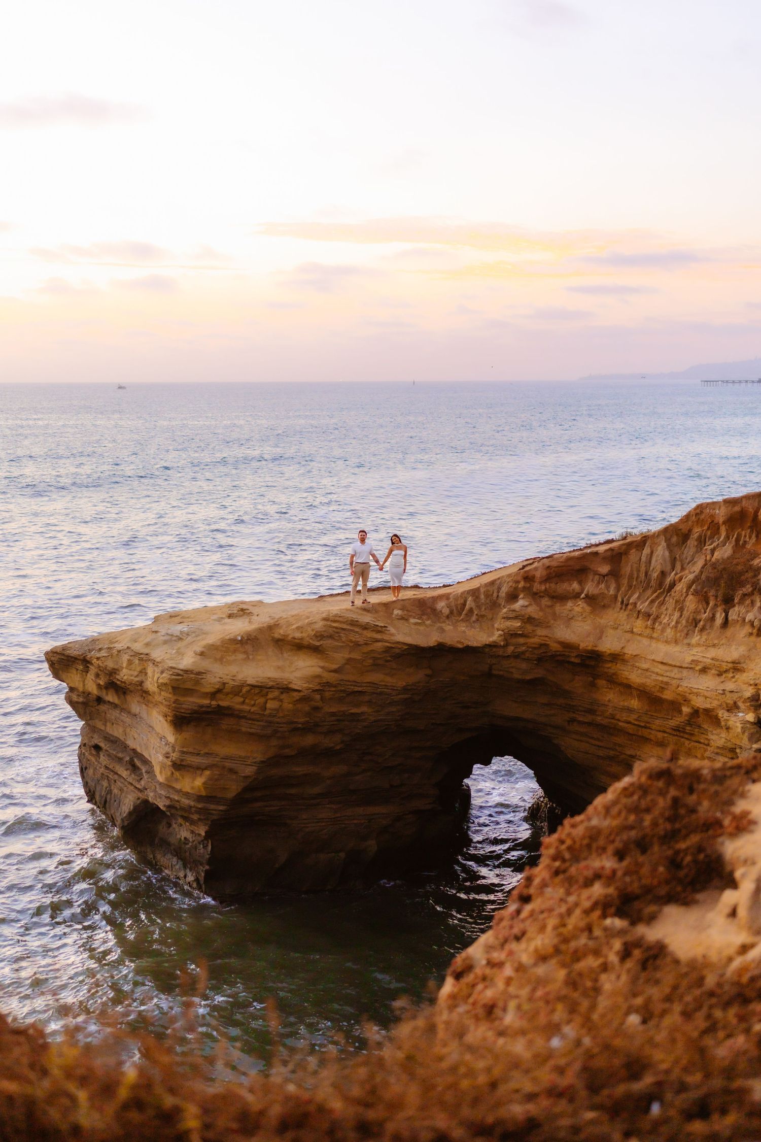 Couple holding hands on dramatic rocky coastal arch Sunset Cliffs at pastel sunset.