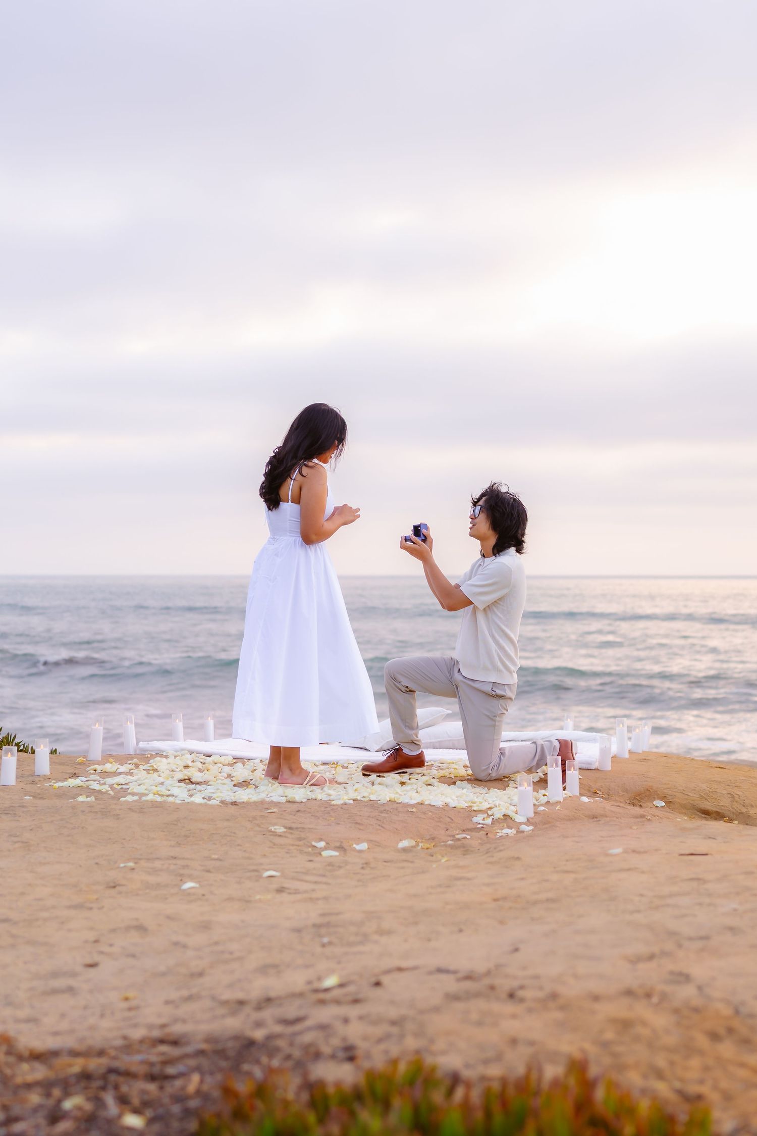 Man proposing on one knee to woman in white dress on sandy cliff with ocean backdrop at dusk.
